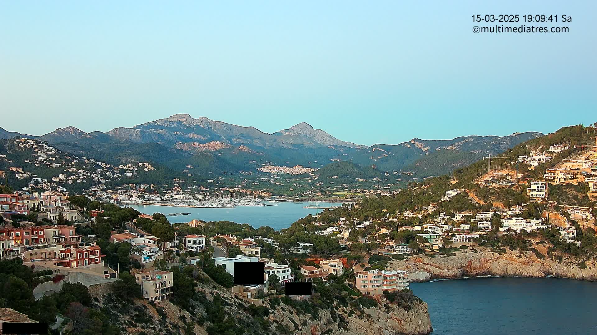 A coastal town nestled in a mountainous region, with a calm bay filled with boats under a clear sky.