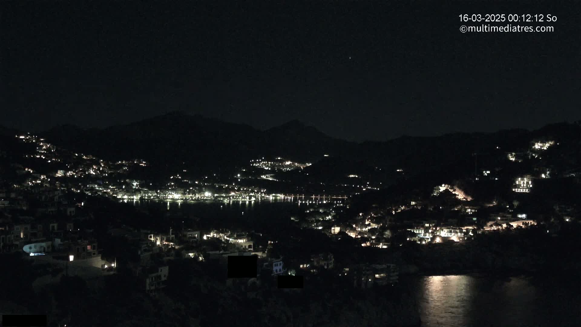 A nighttime view of a coastal town nestled between hills, with scattered lights illuminating the buildings and reflecting on the calm water below.
