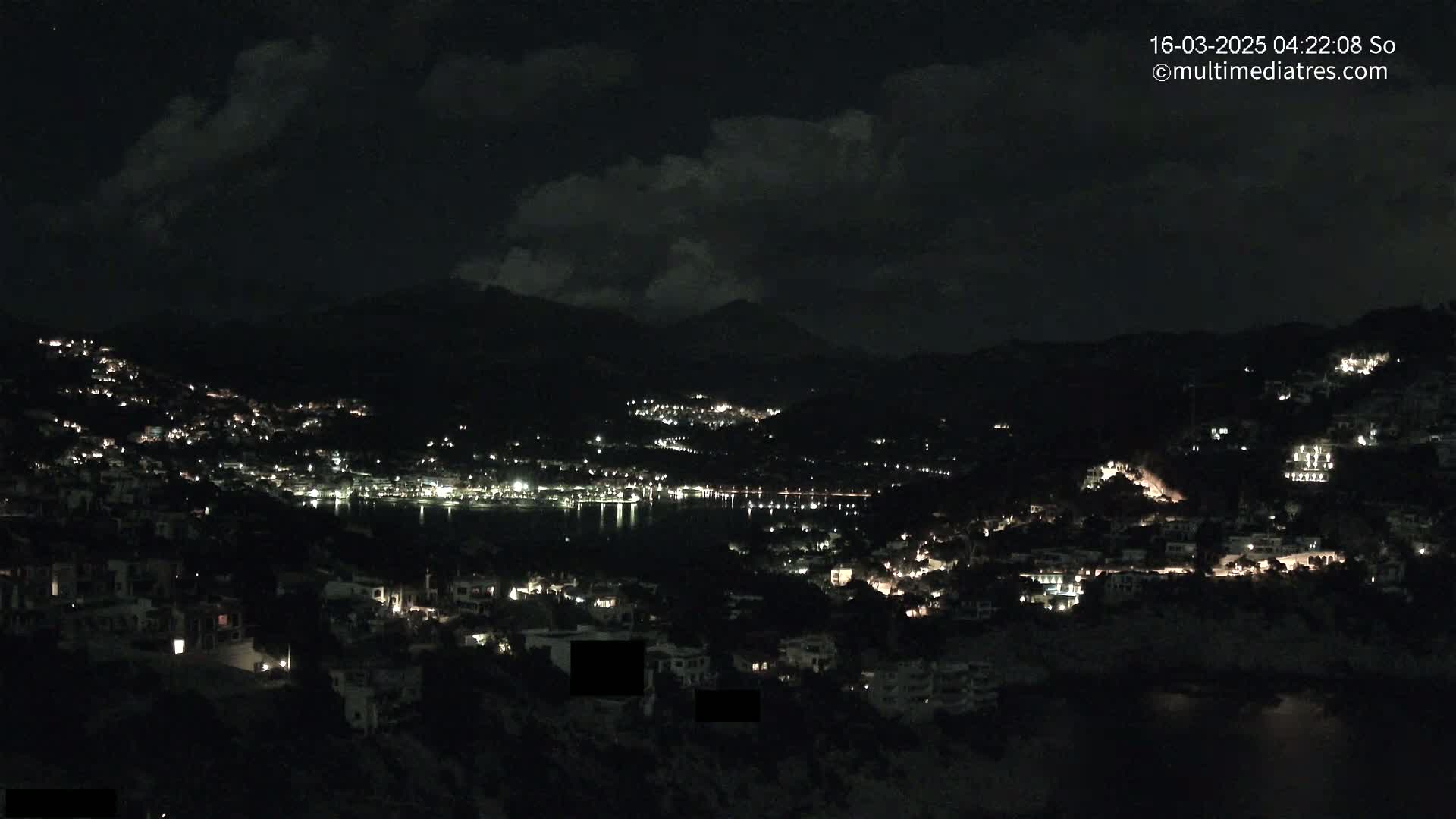 A nighttime view of a coastal town nestled into hills, under a partly cloudy sky, with the lights of buildings visible against the darkness.