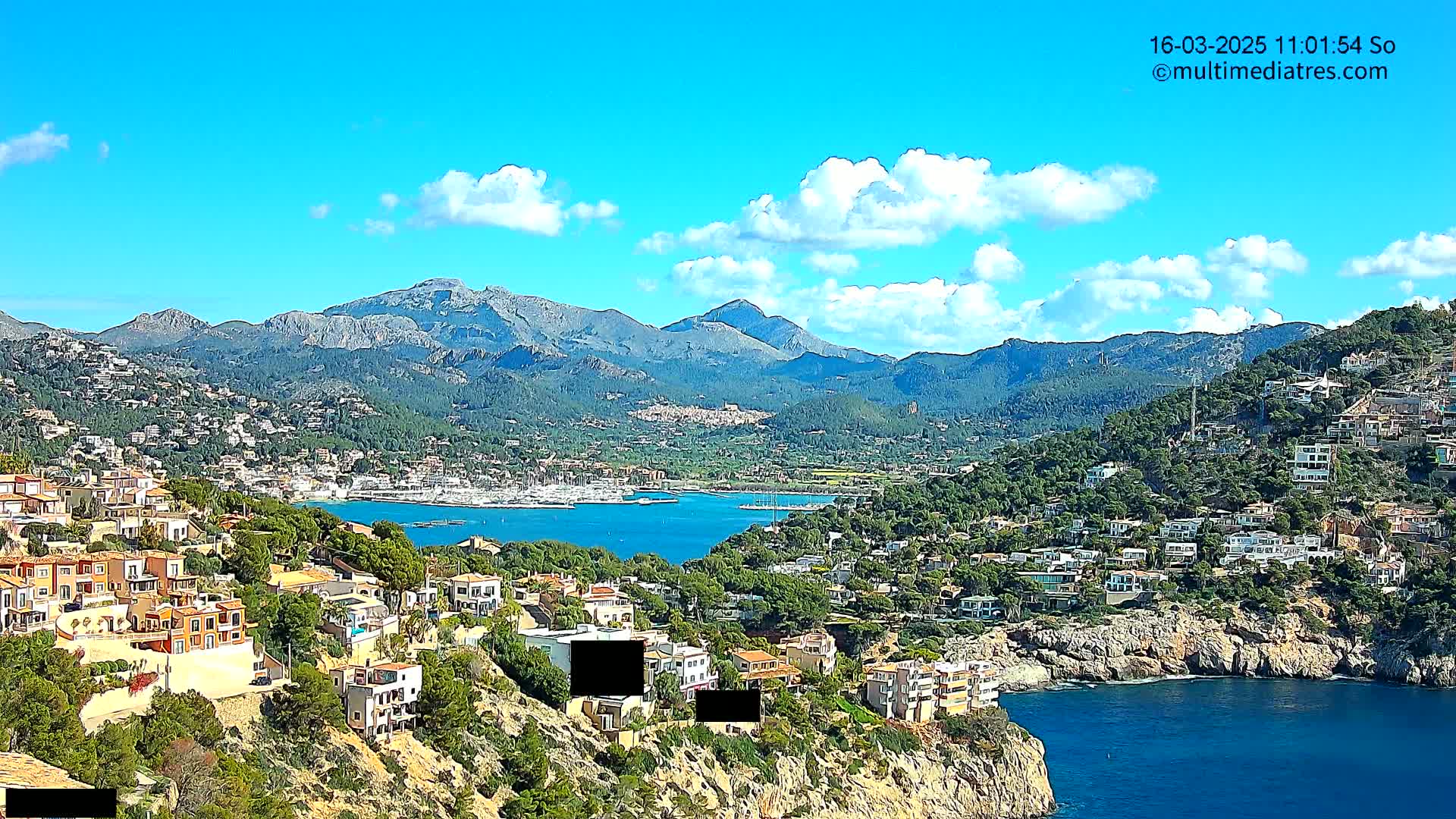 A coastal town nestled on a hillside, overlooking a calm bay with numerous boats and a mountainous backdrop under a mostly sunny sky with scattered clouds.