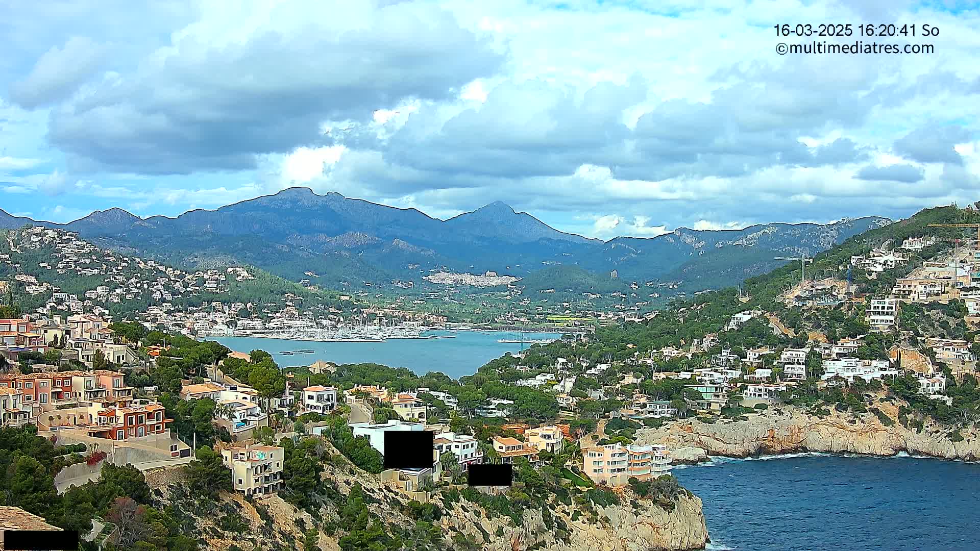 A coastal town nestled on a hillside overlooking a calm bay, with mountains in the background under a partly cloudy sky.