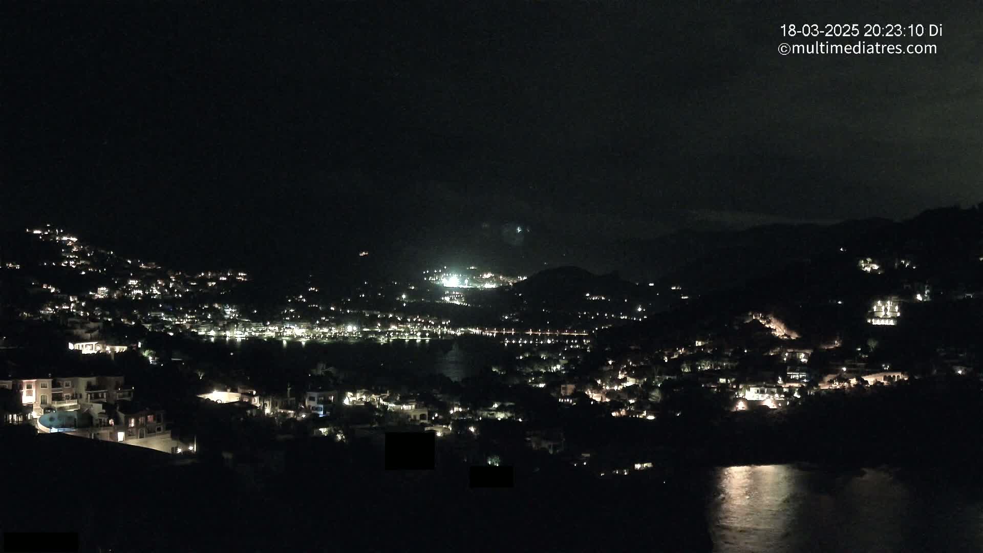 A nighttime view of a coastal town illuminated by many lights, situated on a hillside overlooking dark water.
