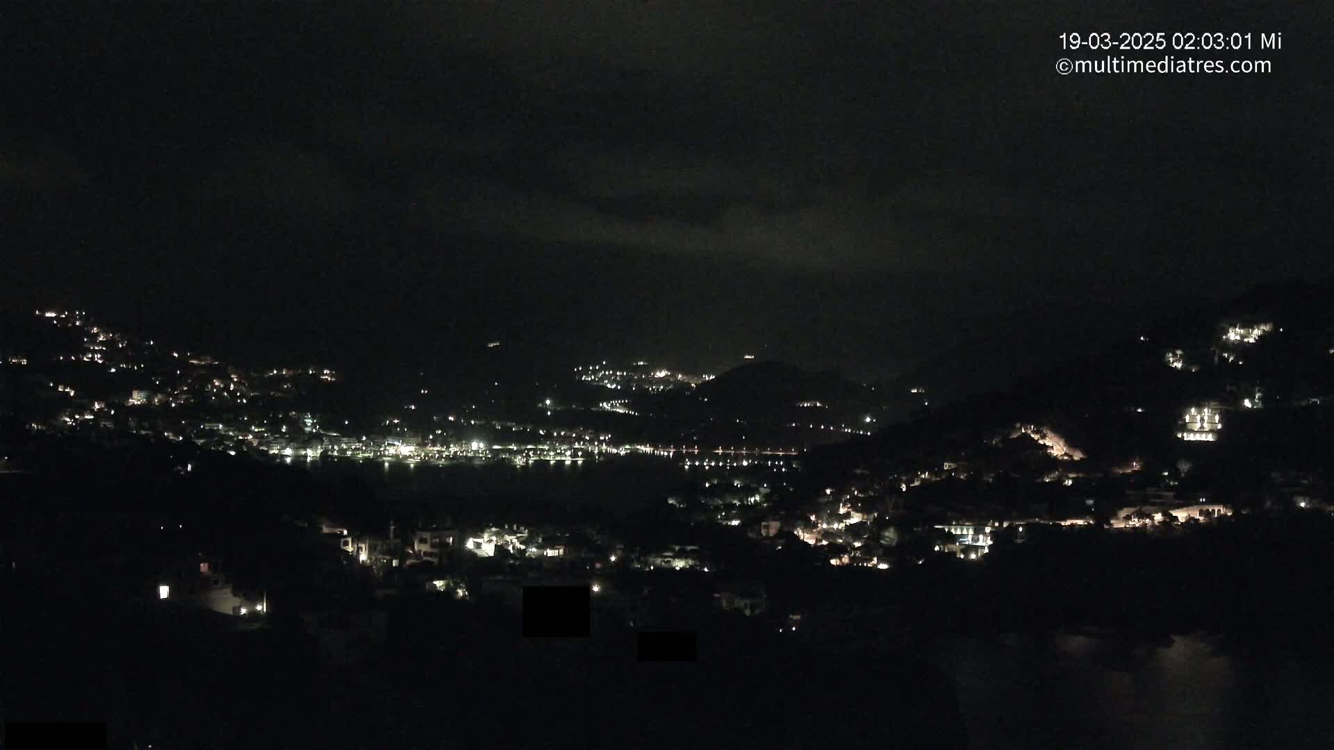 A nighttime view of a coastal town nestled in dark hills, with scattered lights illuminating buildings and a body of water visible in the distance under a mostly cloudy sky.