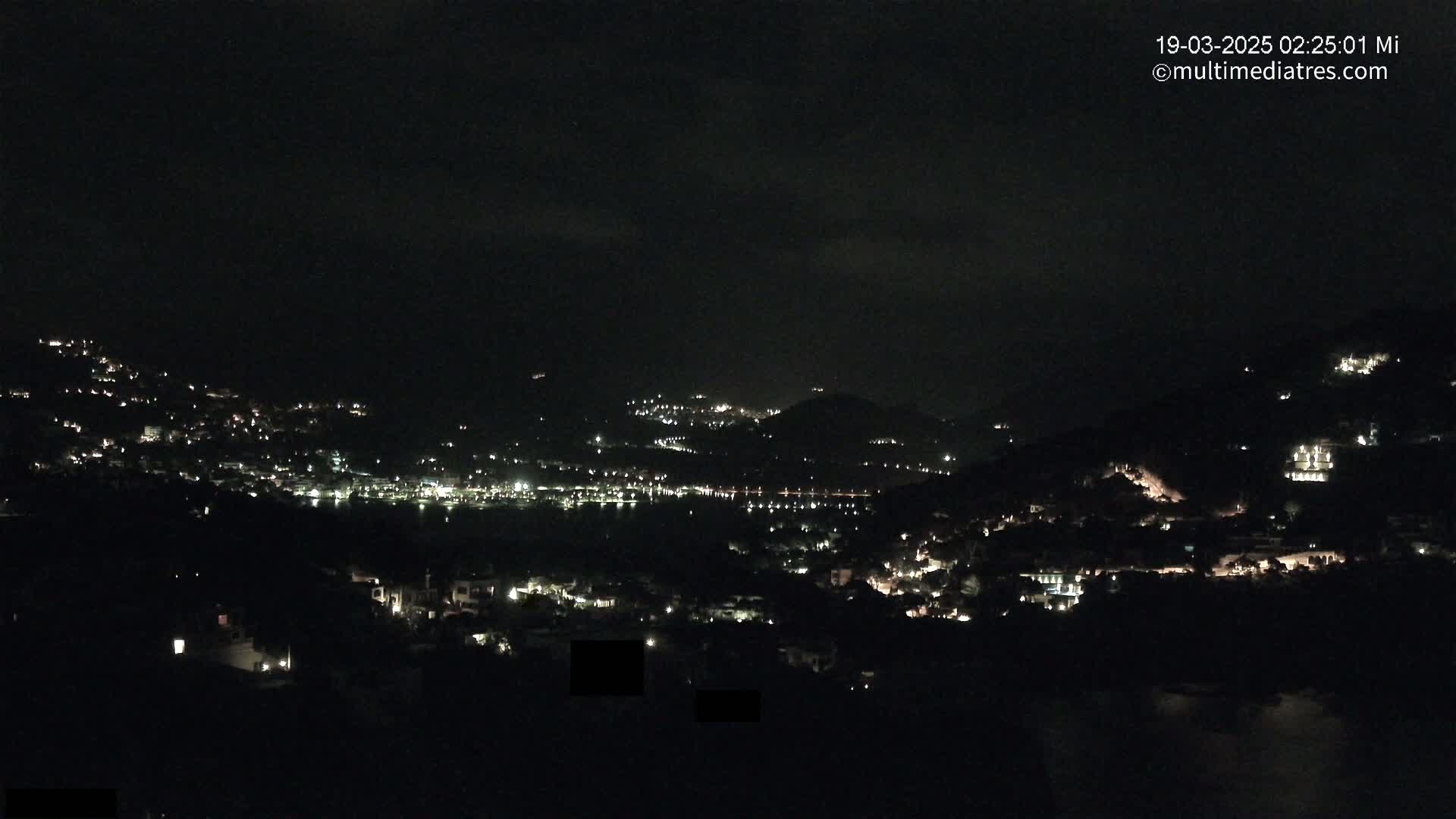 A nighttime view of a coastal town nestled on hillsides, with scattered lights illuminating the buildings and a dark body of water visible in the distance.