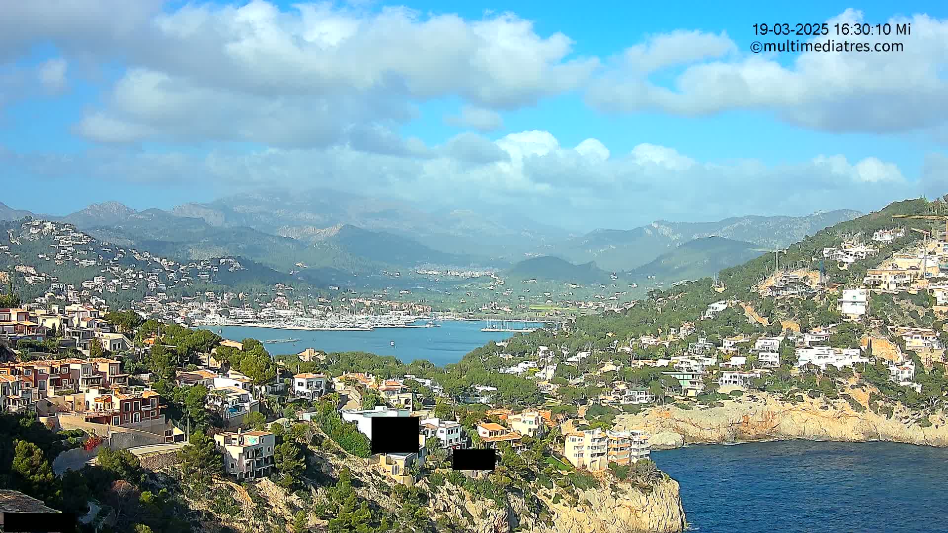 A partly cloudy day reveals a coastal town nestled on a hillside overlooking a calm bay filled with boats, with mountains in the background.