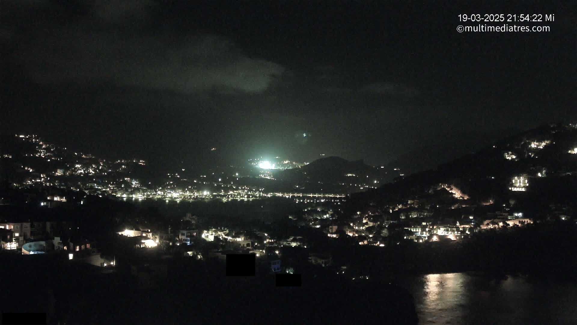 A nighttime view of a coastal town nestled on hillsides, with scattered lights illuminating the buildings and a body of water reflecting light in the foreground under a partly cloudy sky.