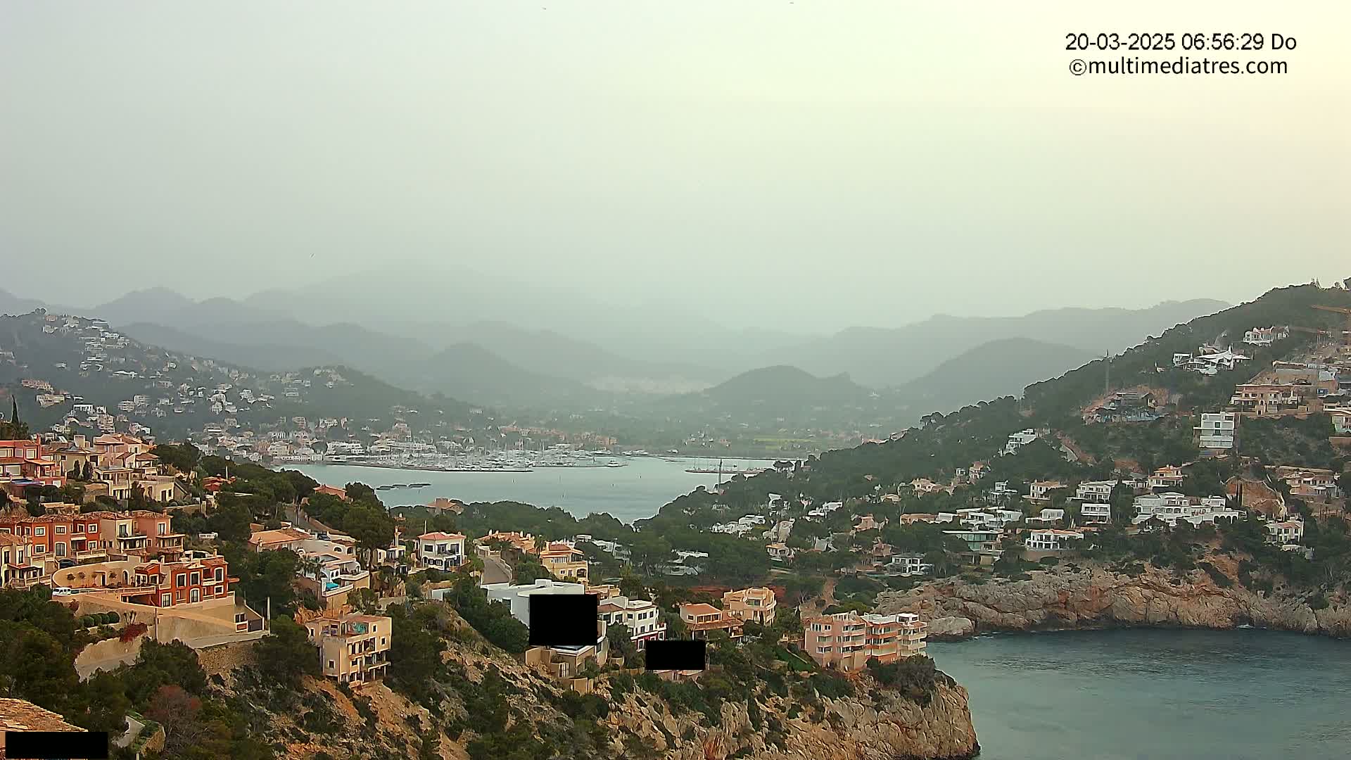 A hazy coastal scene shows a bay with a marina, surrounded by hills dotted with houses, under a muted sky.
