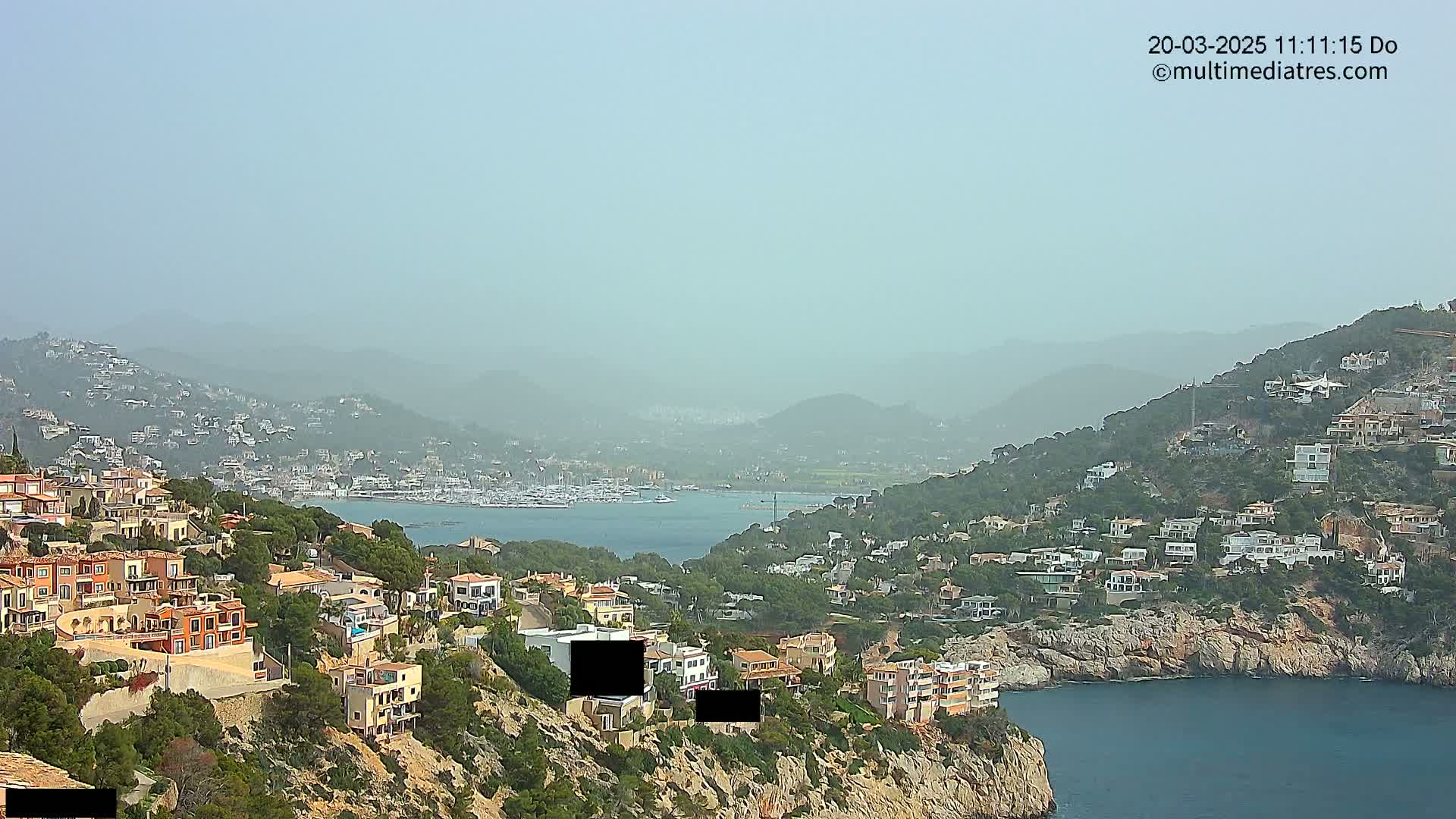 A hazy coastal view shows a hillside town overlooking a calm bay with many boats, all situated against a backdrop of distant mountains.