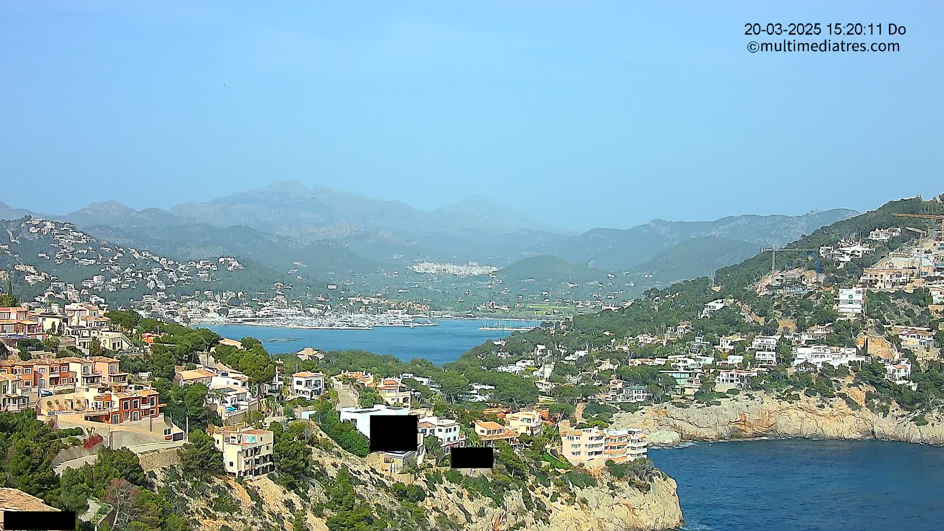 A clear, sunny day overlooks a coastal town nestled between a mountain range and a calm bay dotted with sailboats and houses.