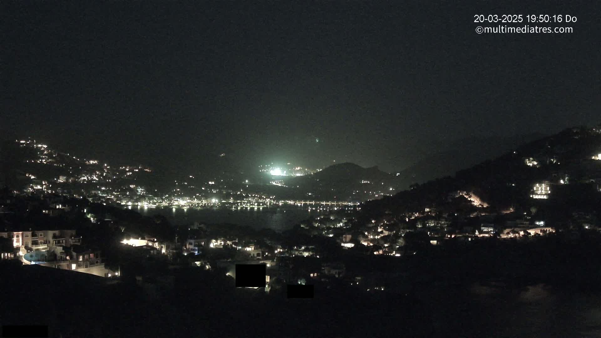 A nighttime view of a coastal town nestled on hillsides, with numerous lights illuminating buildings and streets along a dark body of water under a clear sky.