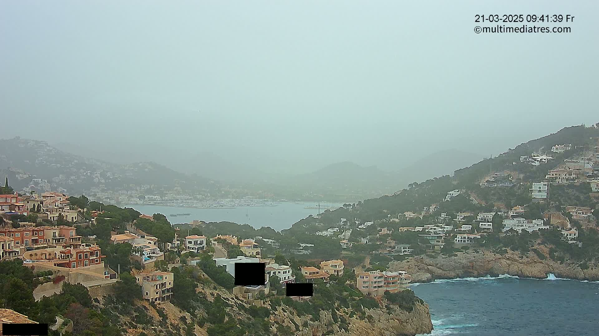 A hazy coastal scene shows a hillside dotted with houses overlooking a calm bay with boats, under a dusty sky.
