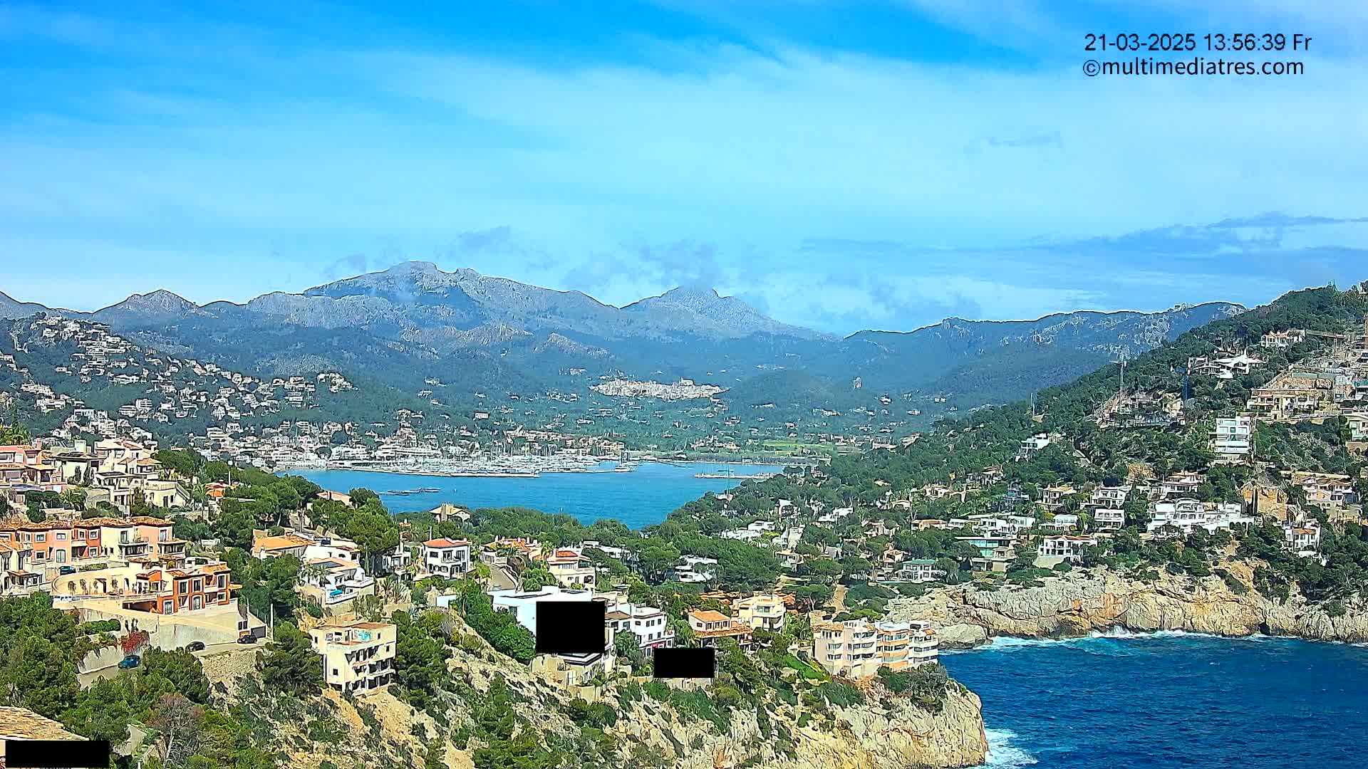 A coastal town nestled on a hillside overlooking a calm bay and distant mountains under a clear blue sky.