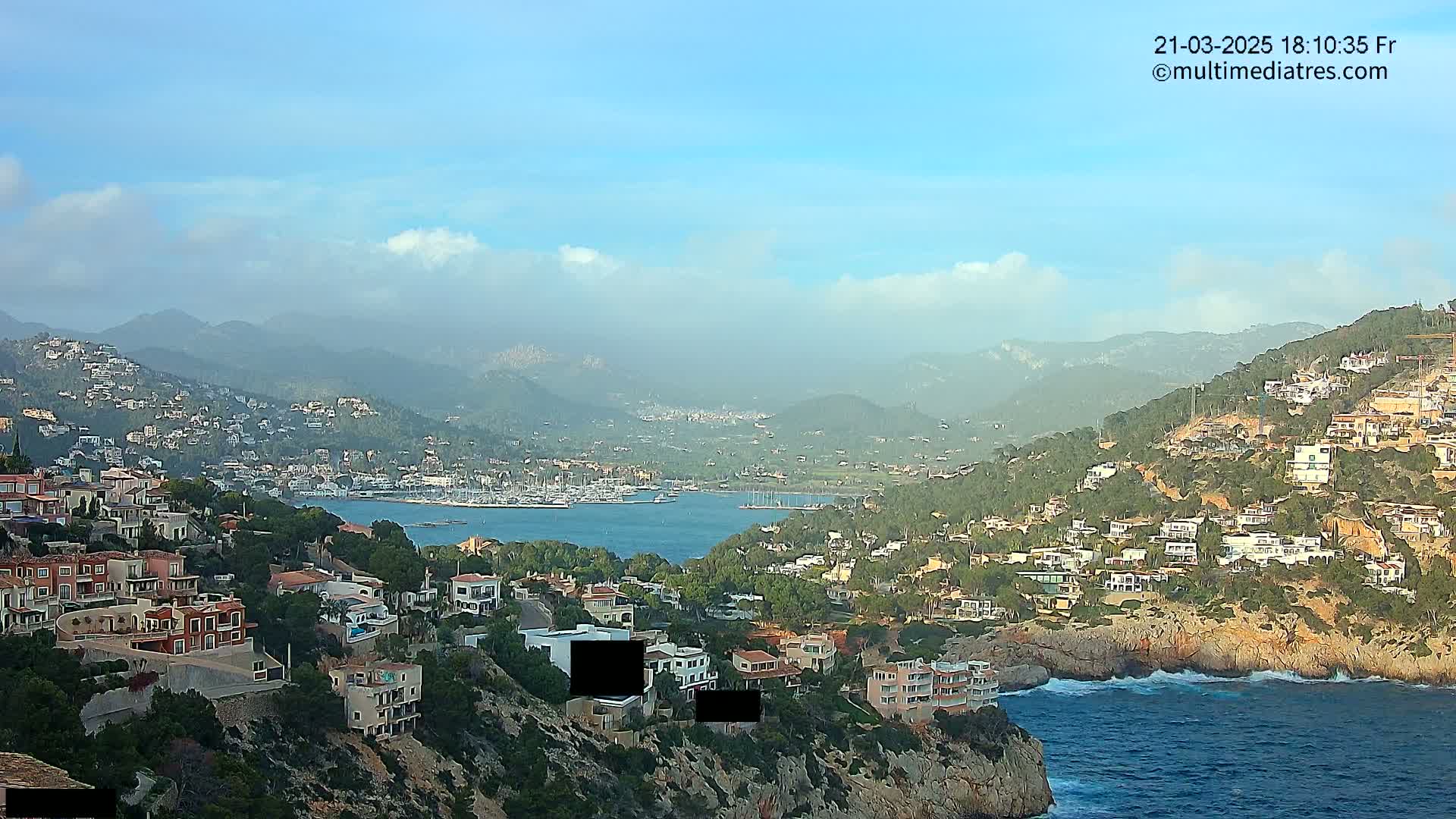 A coastal town nestled in a hillside overlooking a calm bay filled with boats, under a mostly clear sky with some clouds.