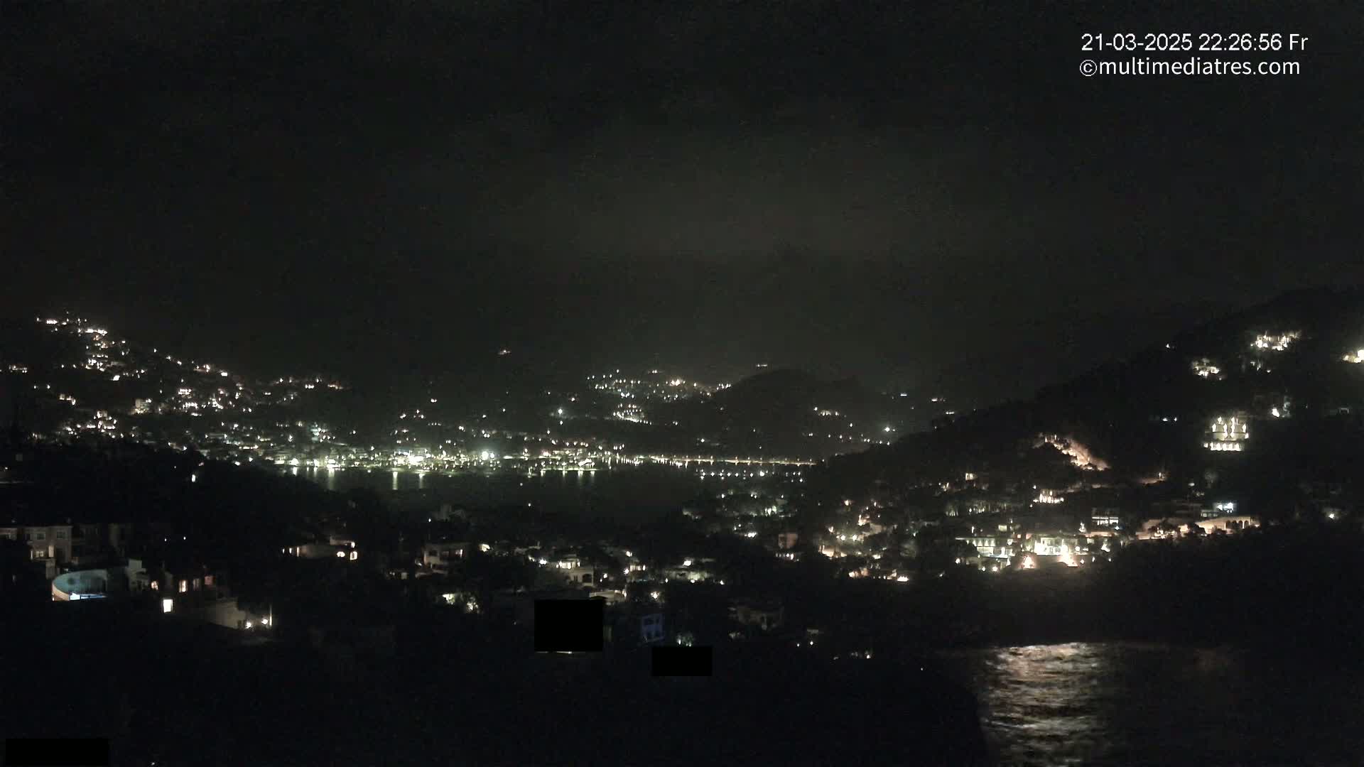A nighttime view of a coastal town nestled on hillsides, with scattered lights illuminating the buildings and a dark, calm body of water reflecting some light.