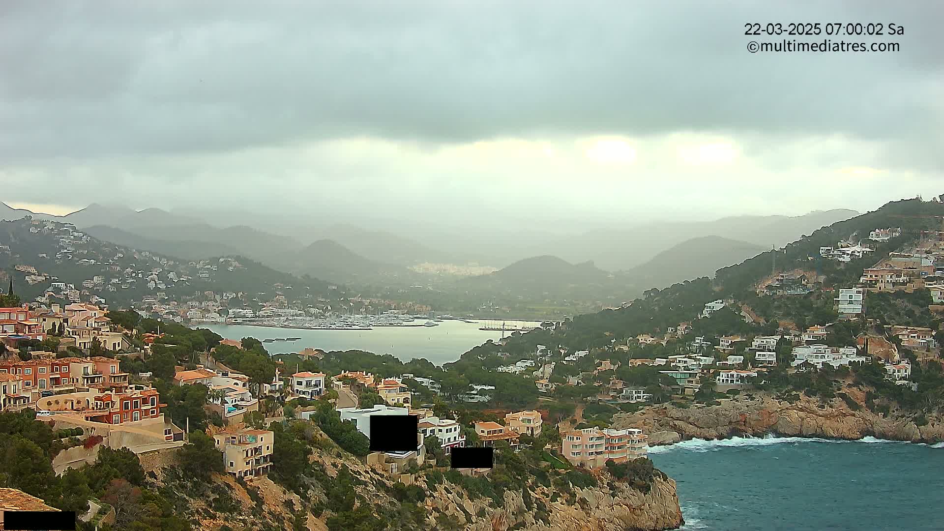 A coastal town nestled in a hillside overlooking a calm bay with many boats, under an overcast sky.