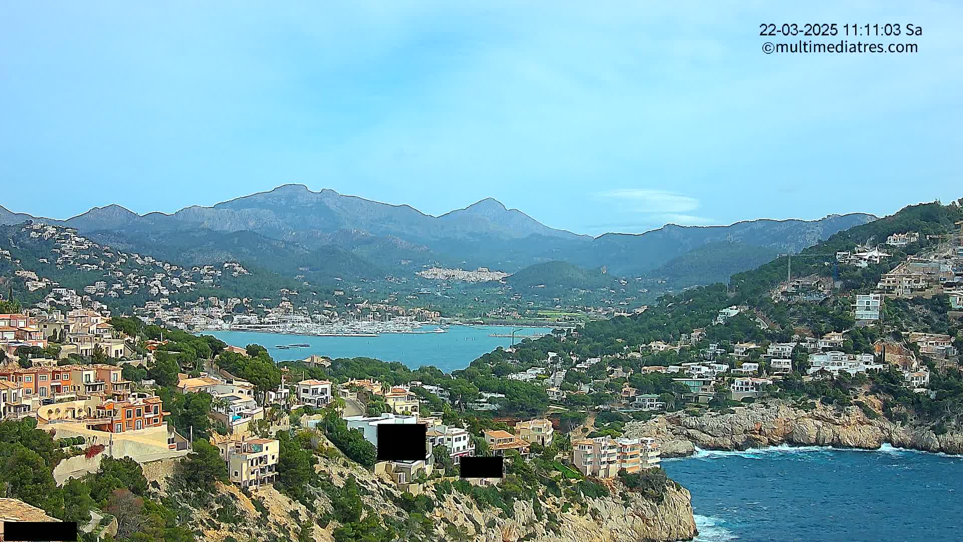A coastal town nestled on a hillside overlooking a calm bay with a marina and mountains in the background under a mostly clear sky.