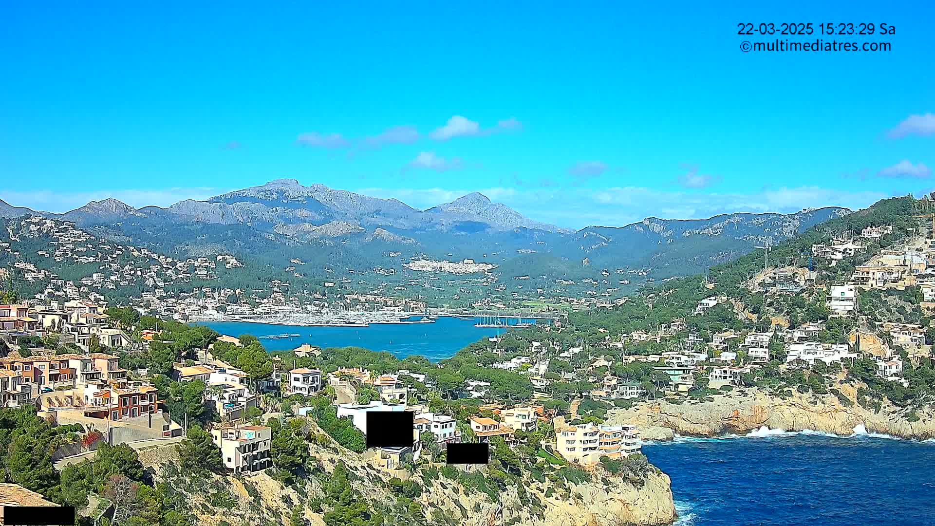 A coastal town nestled against a mountain range under a clear blue sky, featuring a calm bay with numerous boats.