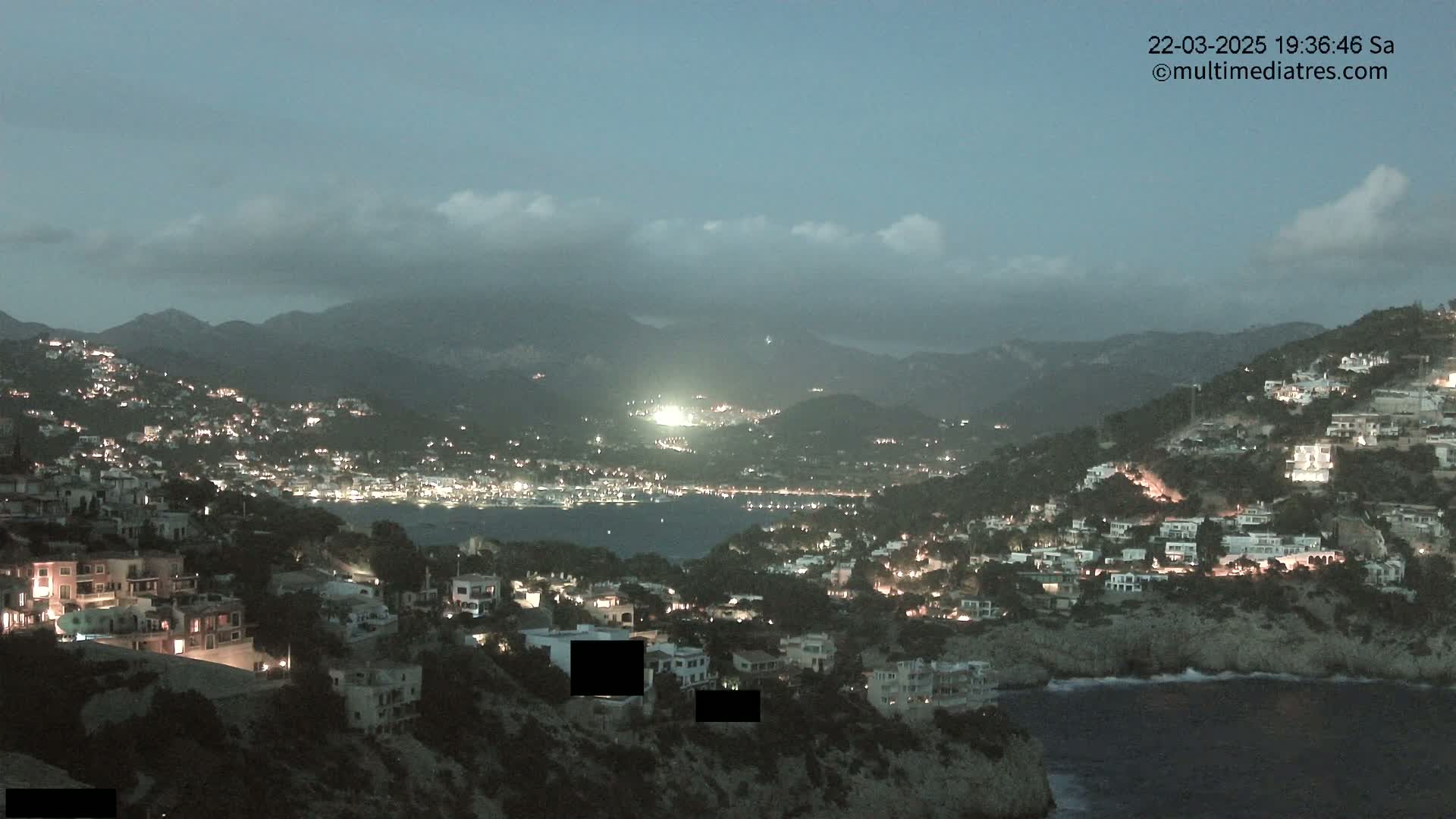 A nighttime coastal town illuminated with lights, nestled among dark hills under a partly cloudy sky.