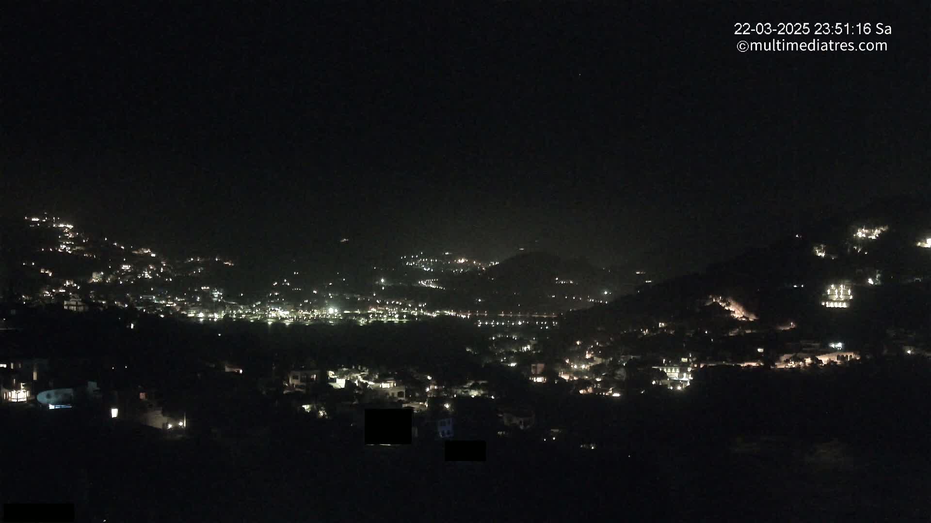 A nighttime view of a town nestled on hillsides, illuminated by scattered lights under a clear, dark sky.