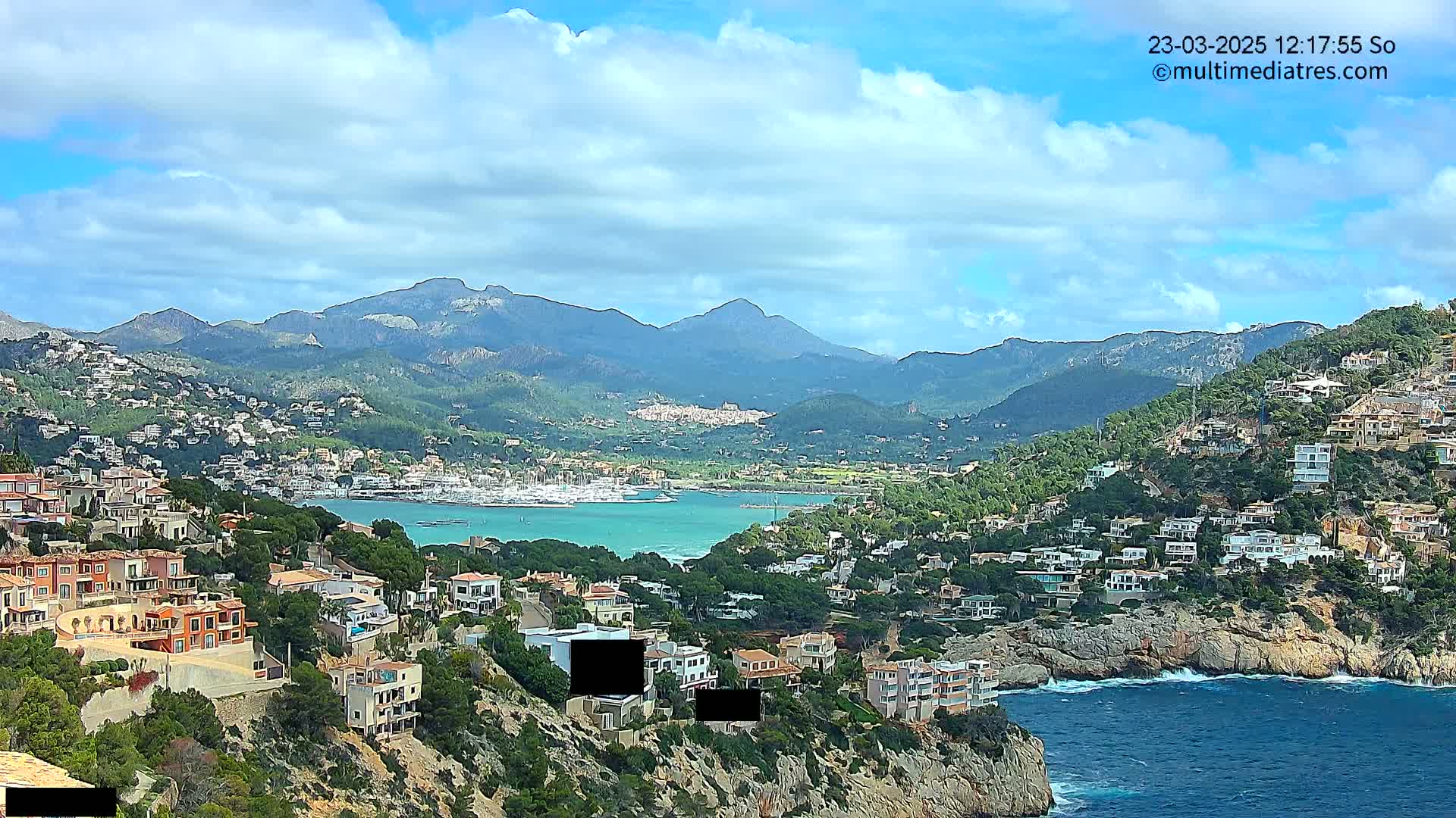 A coastal town nestled on a hillside overlooking a calm, turquoise bay, with mountains in the background under a partly cloudy sky.