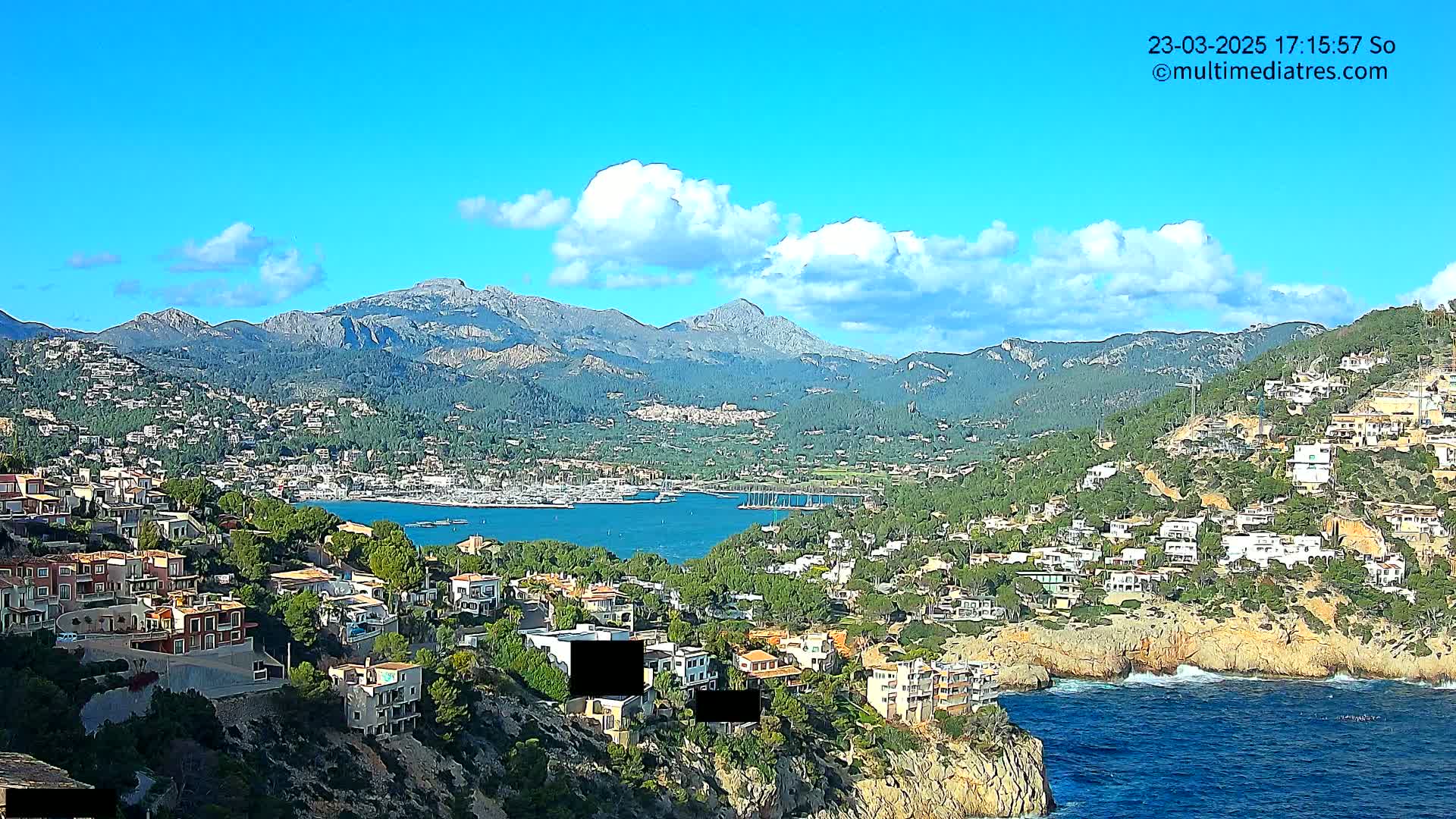 A coastal town nestled in a mountainous region overlooks a calm bay with boats, under a mostly sunny sky with a few clouds.