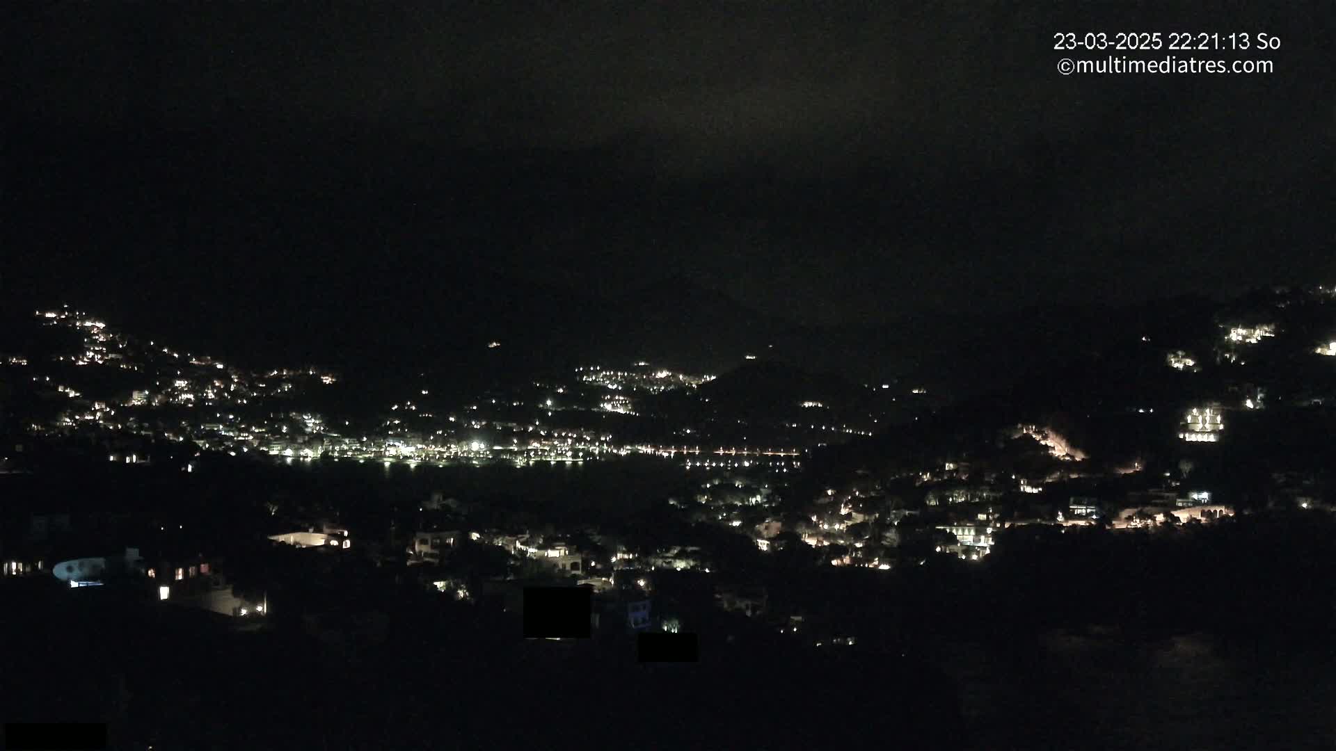 A nighttime view of a town nestled on a hillside, illuminated by scattered lights under a dark, cloudy sky.