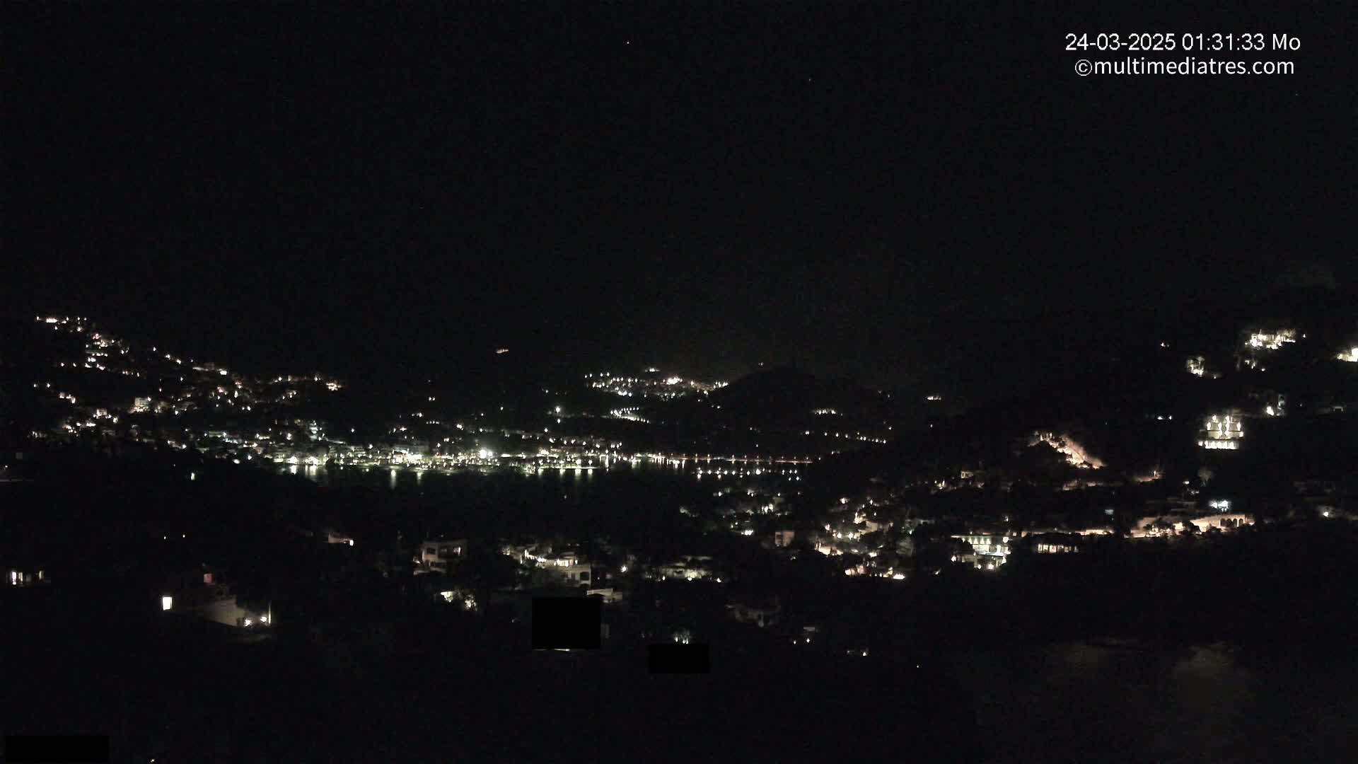 A nighttime, long-range view shows a coastal town illuminated by scattered lights, nestled on hillsides beside a dark body of water under a clear, starless sky.