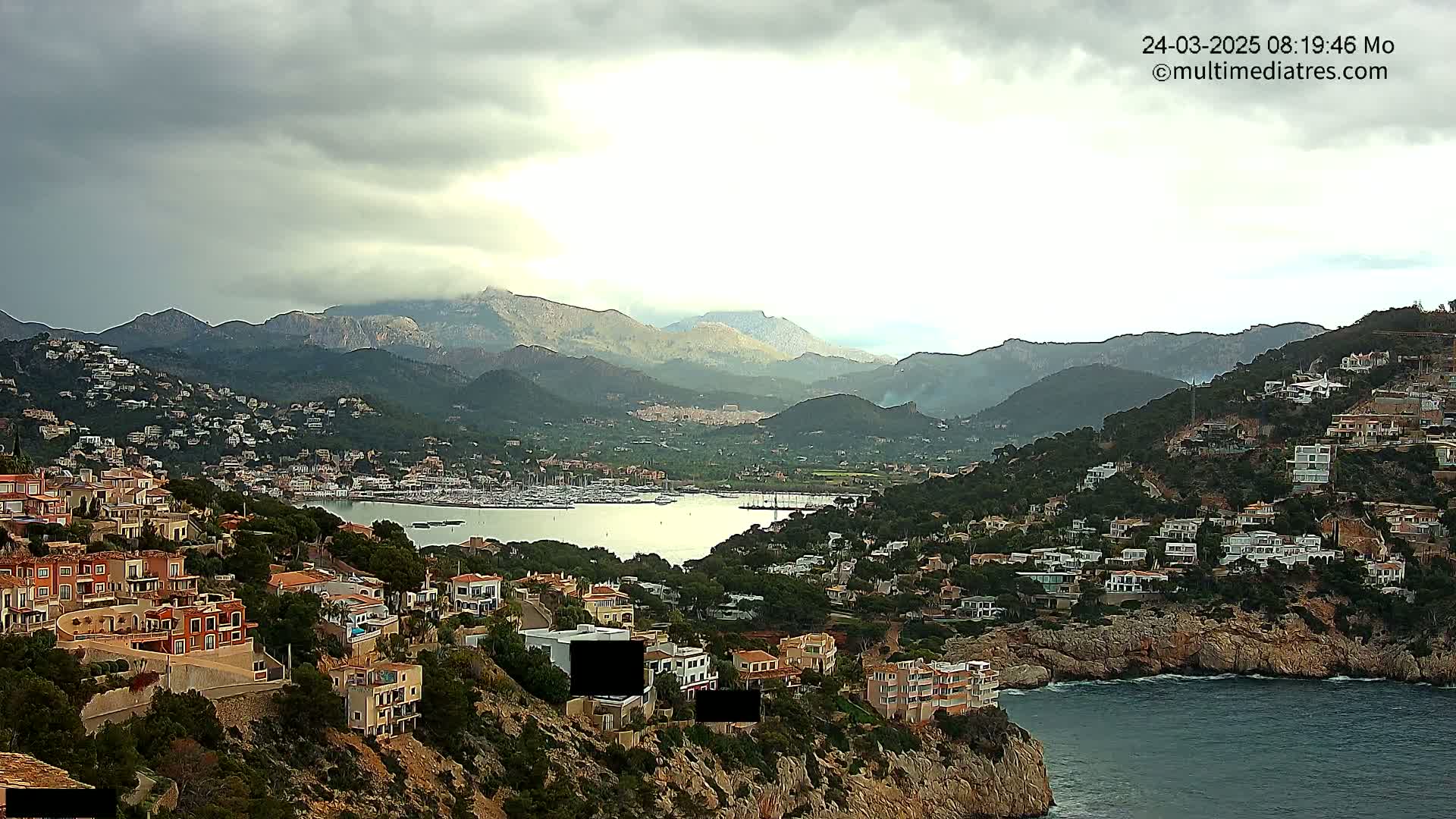 A coastal town nestled in a mountainous region under a cloudy sky overlooks a calm bay filled with boats.
