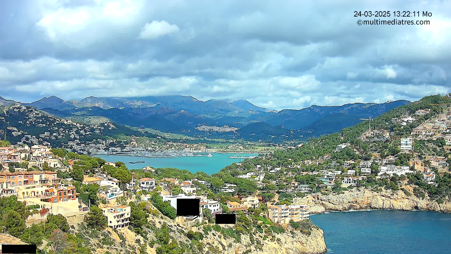 A coastal town nestled on a hillside overlooking a calm bay and distant mountains under a partly cloudy sky.