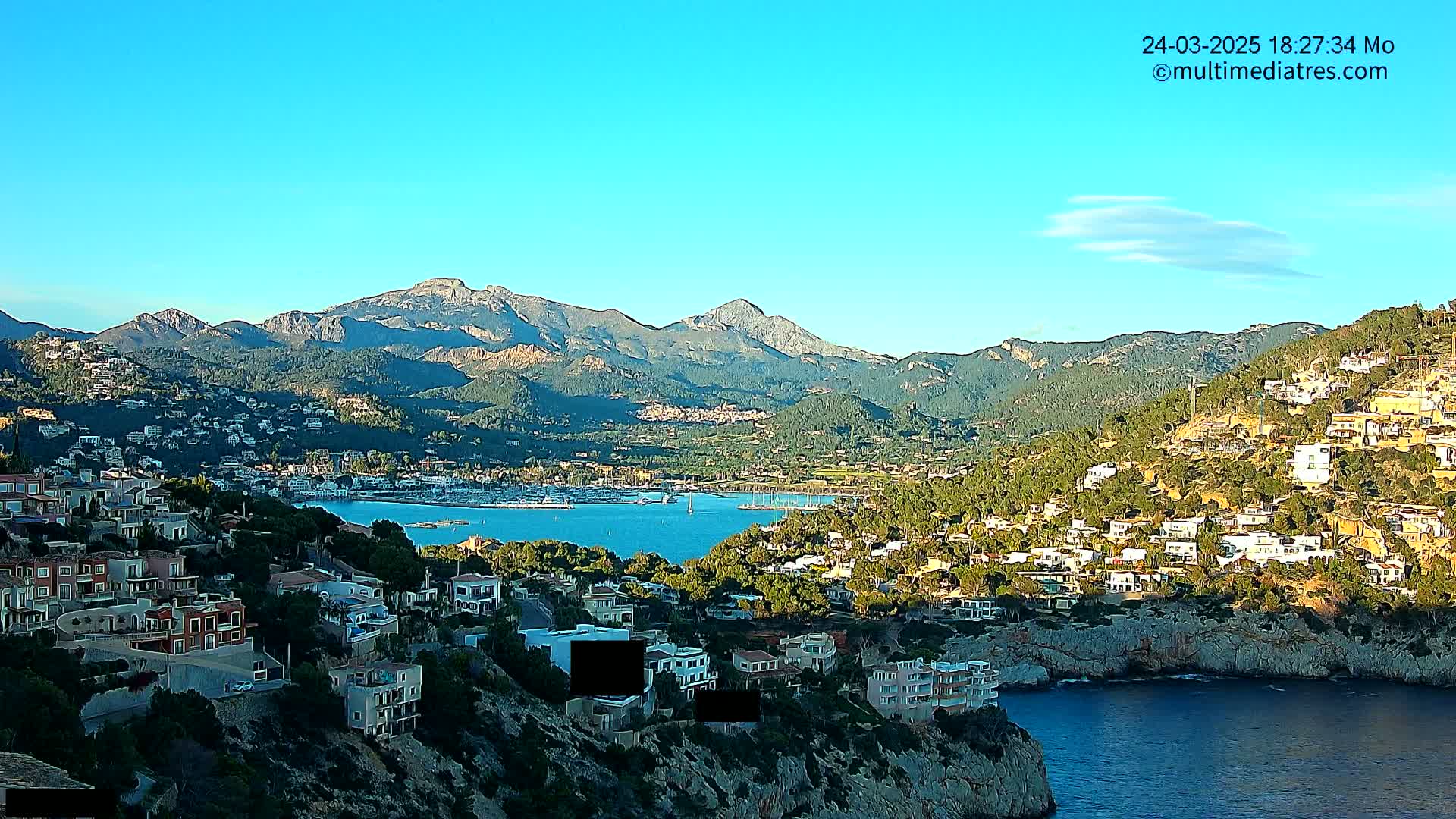 A coastal town nestled on a hillside overlooking a calm bay, with mountains in the background under a clear blue sky.