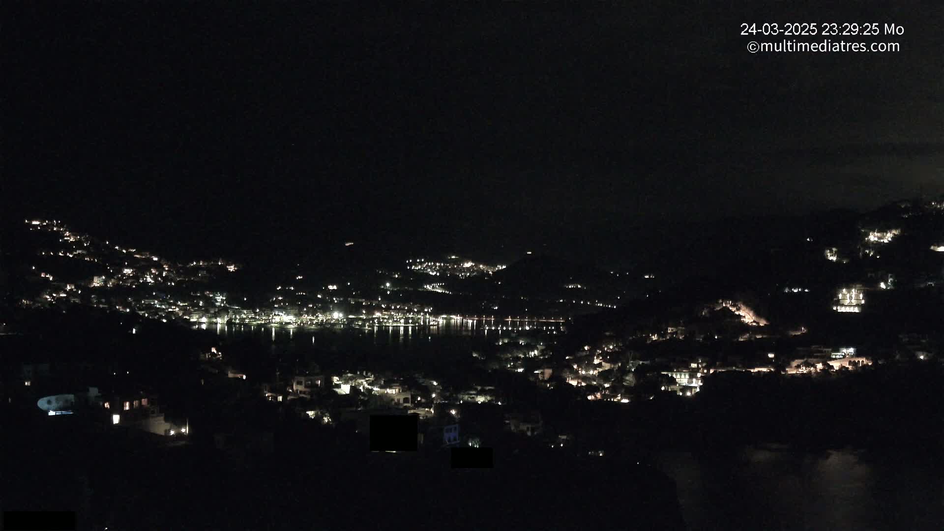 A nighttime view of a town nestled on hillsides surrounding a calm body of water.
