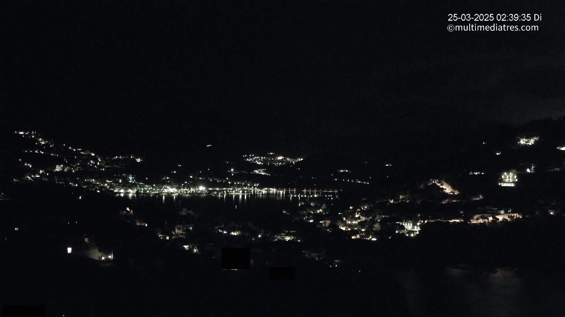 A nighttime view of a town nestled on a hillside, its lights reflecting on a calm body of water below.
