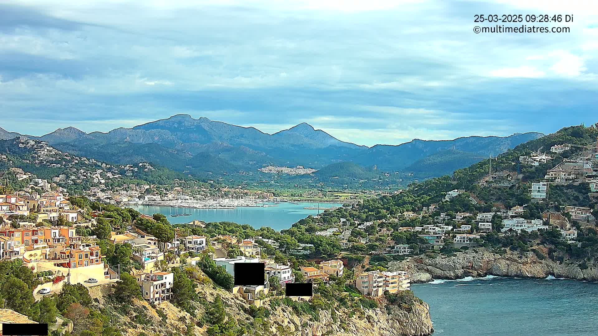 A coastal town nestled into a hillside overlooking a calm bay filled with boats, with mountains in the background under a partly cloudy sky.