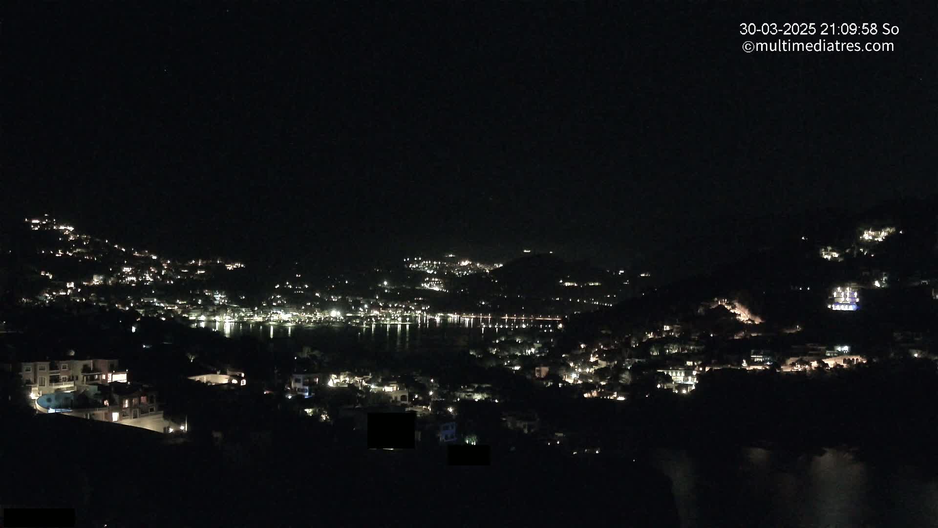 A nighttime view shows a town's lights spread across hillsides and along a dark body of water.
