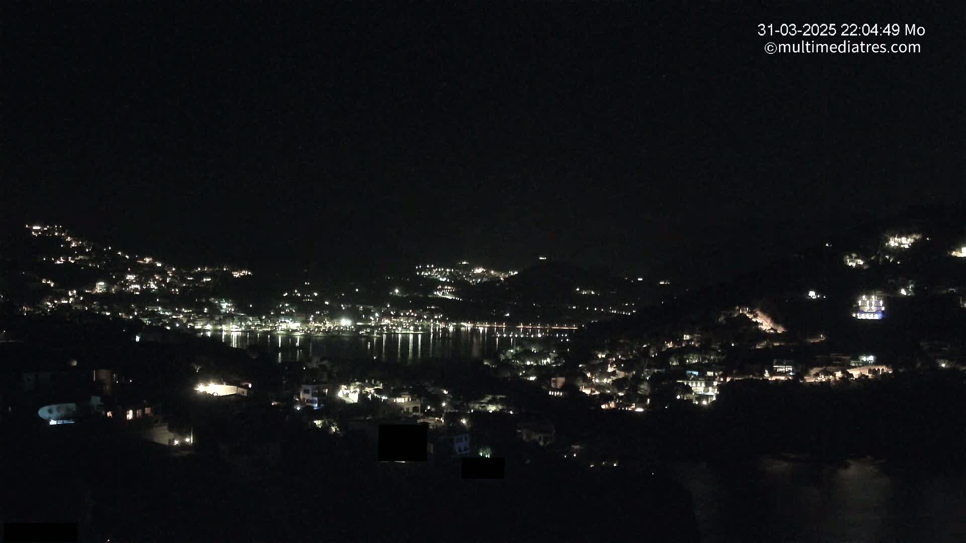 A nighttime view of a coastal town illuminated by scattered lights, with a calm body of water reflecting the lights.