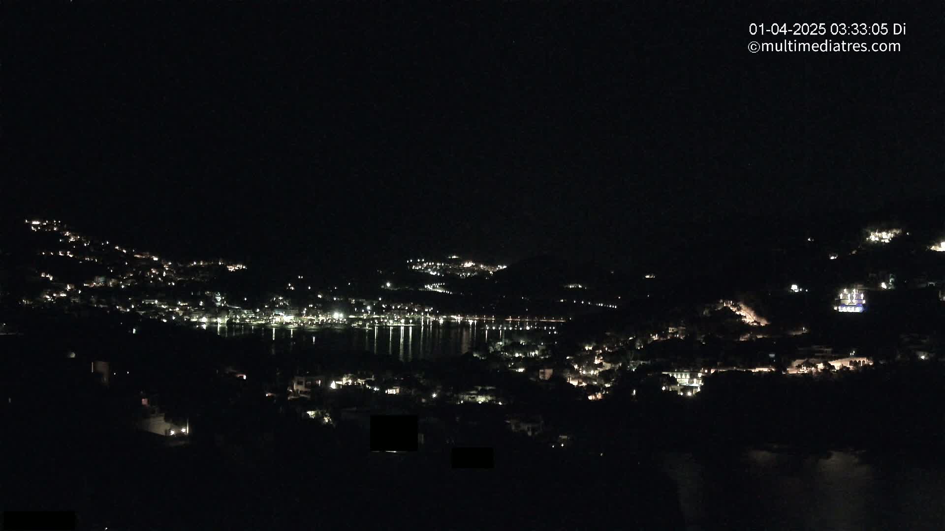 A nighttime view of a coastal town nestled on hillsides, with its lights reflected on a calm body of water under a clear, dark sky.