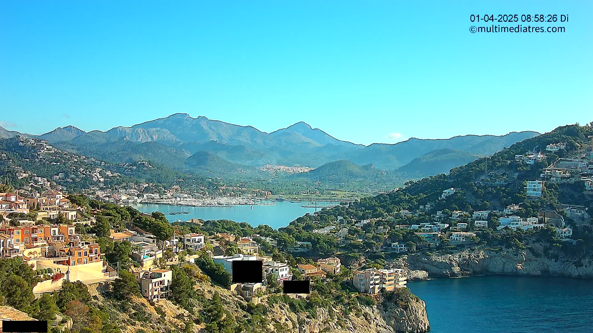 A coastal town nestled in a mountainous region on a clear, sunny day, with a calm bay filled with boats in the foreground.