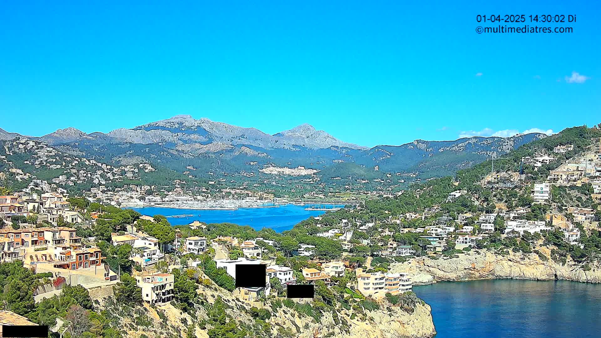 A coastal town nestled in a mountainous region under a clear blue sky, with a calm body of water featuring a marina.