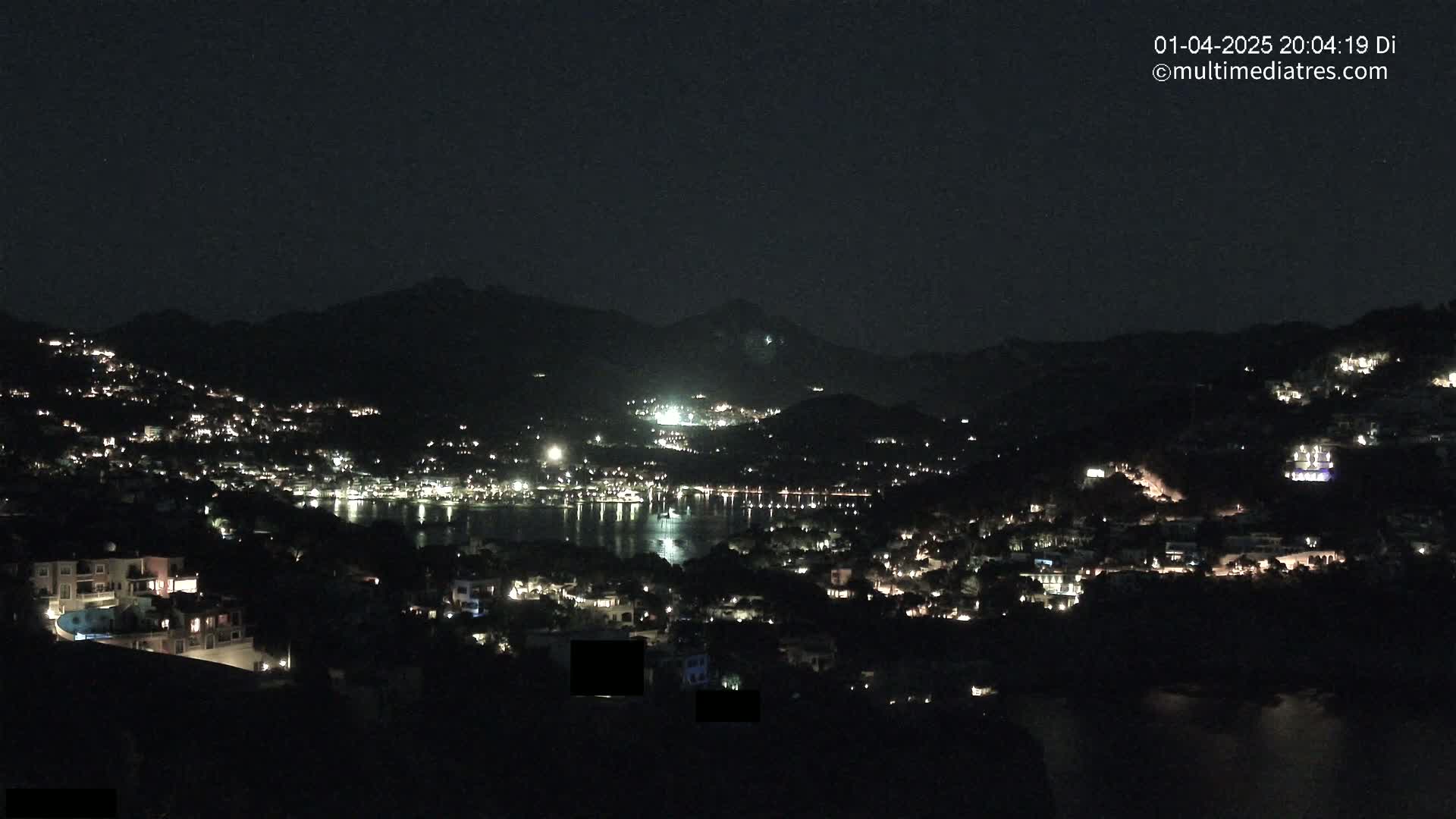 A nighttime view of a town nestled in hills overlooking a dark body of water, illuminated by scattered lights.