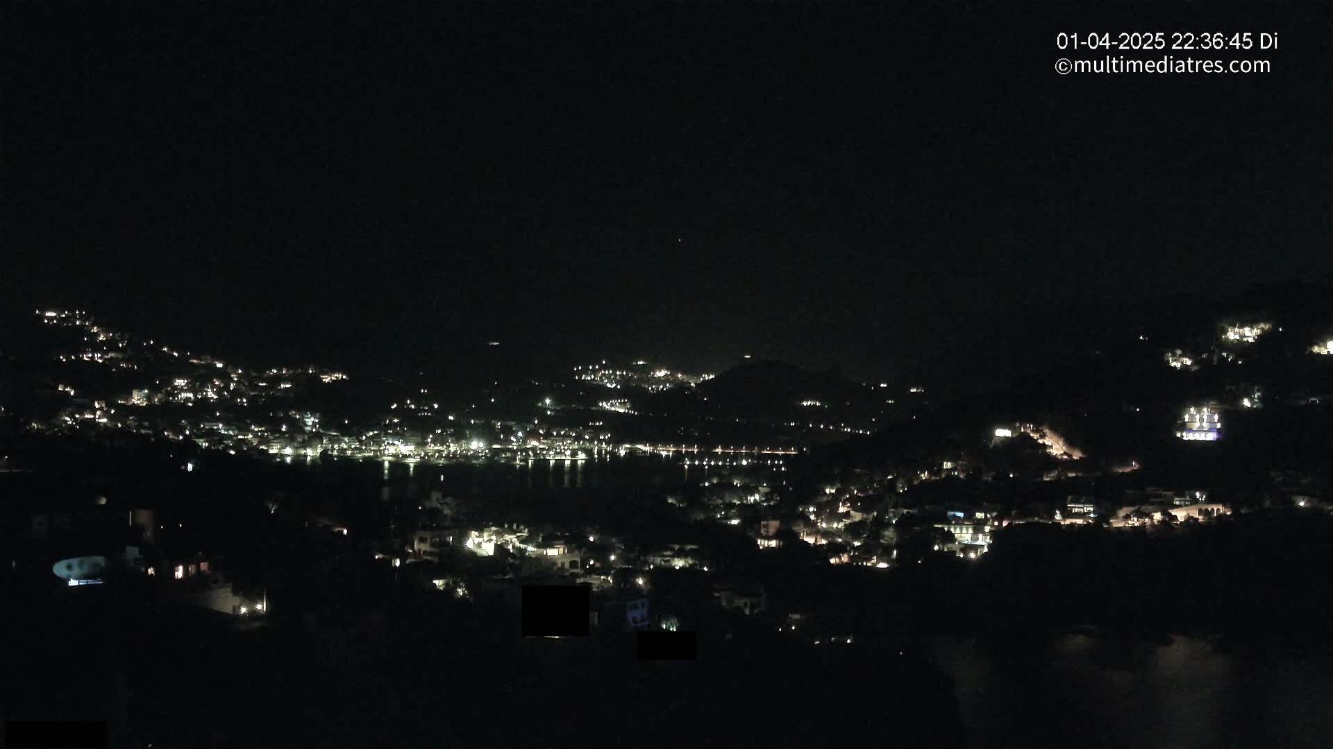 A nighttime view of a coastal town illuminated by scattered lights, nestled on hillsides overlooking a dark body of water under a clear, dark sky.