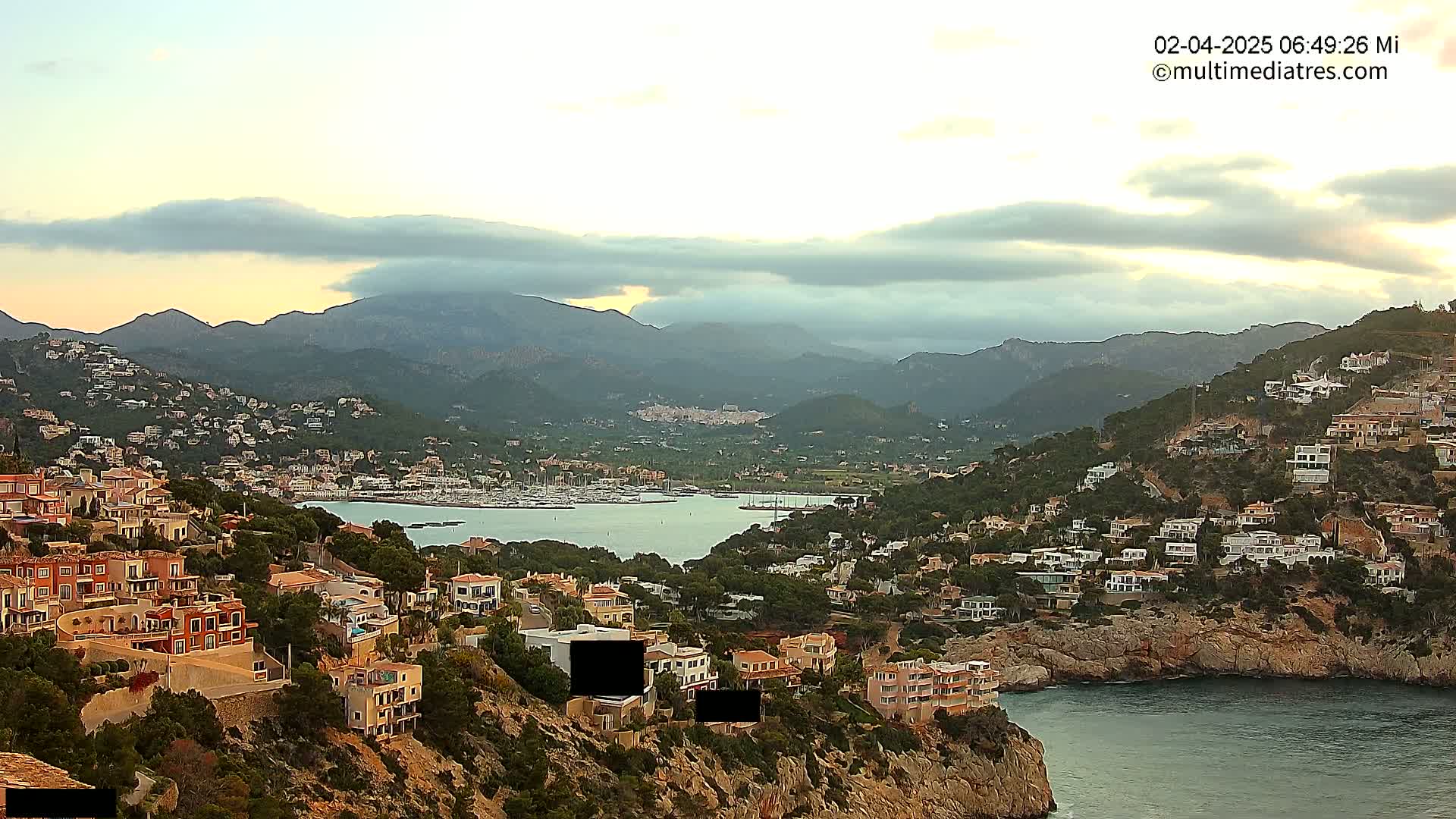 A coastal town nestled in a mountainous region overlooks a calm bay filled with boats under a partly cloudy sky.