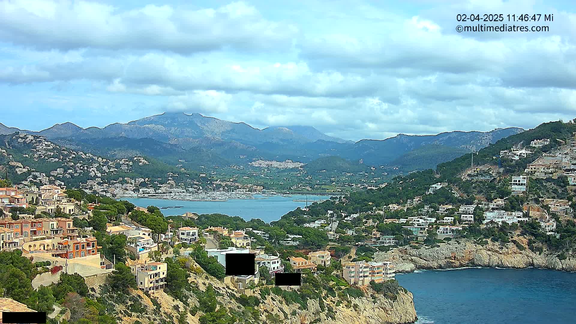 A coastal town nestled into a hillside overlooking a calm bay and a mountain range under a partly cloudy sky.