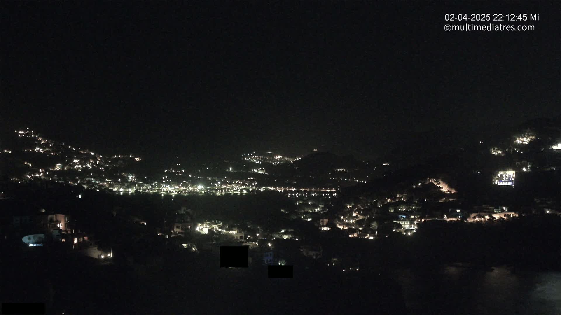A nighttime view of a hillside town illuminated by scattered lights, under a clear, dark sky.