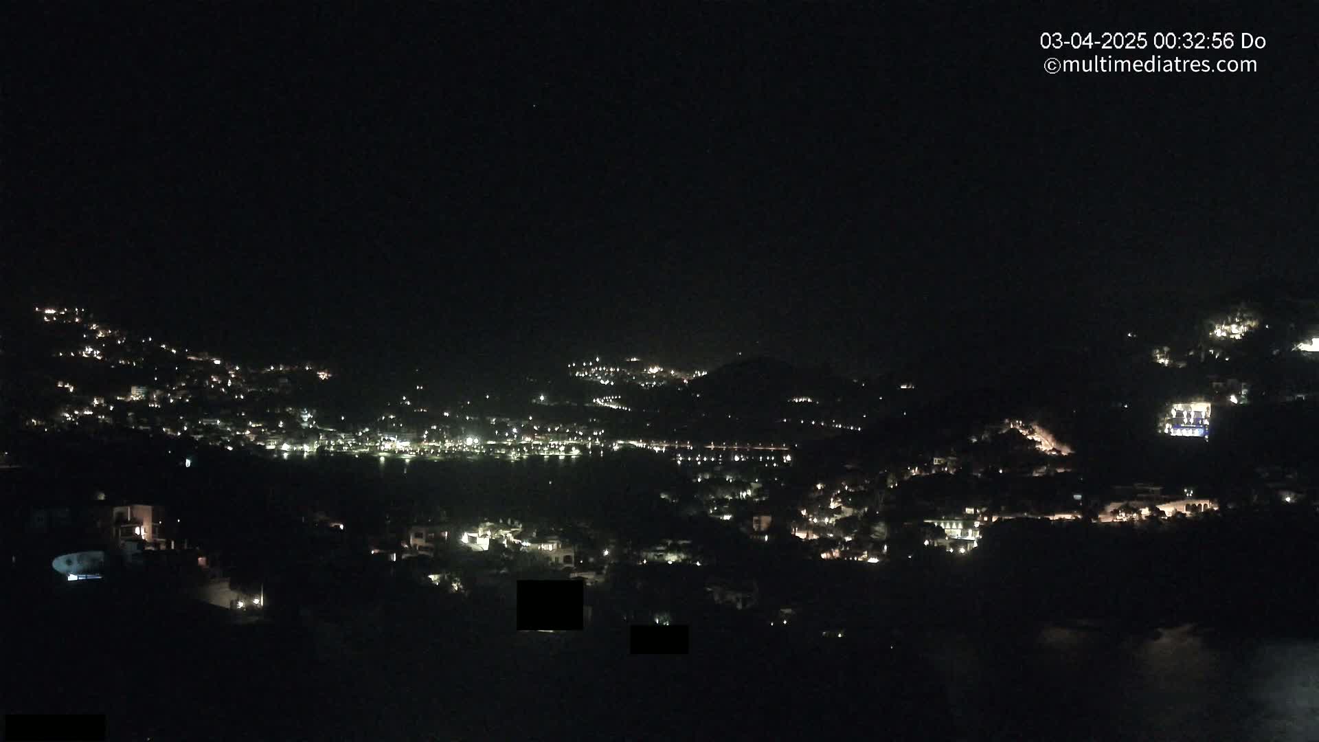 A nighttime view of a hillside town with scattered lights, overlooking a body of water also dotted with lights.