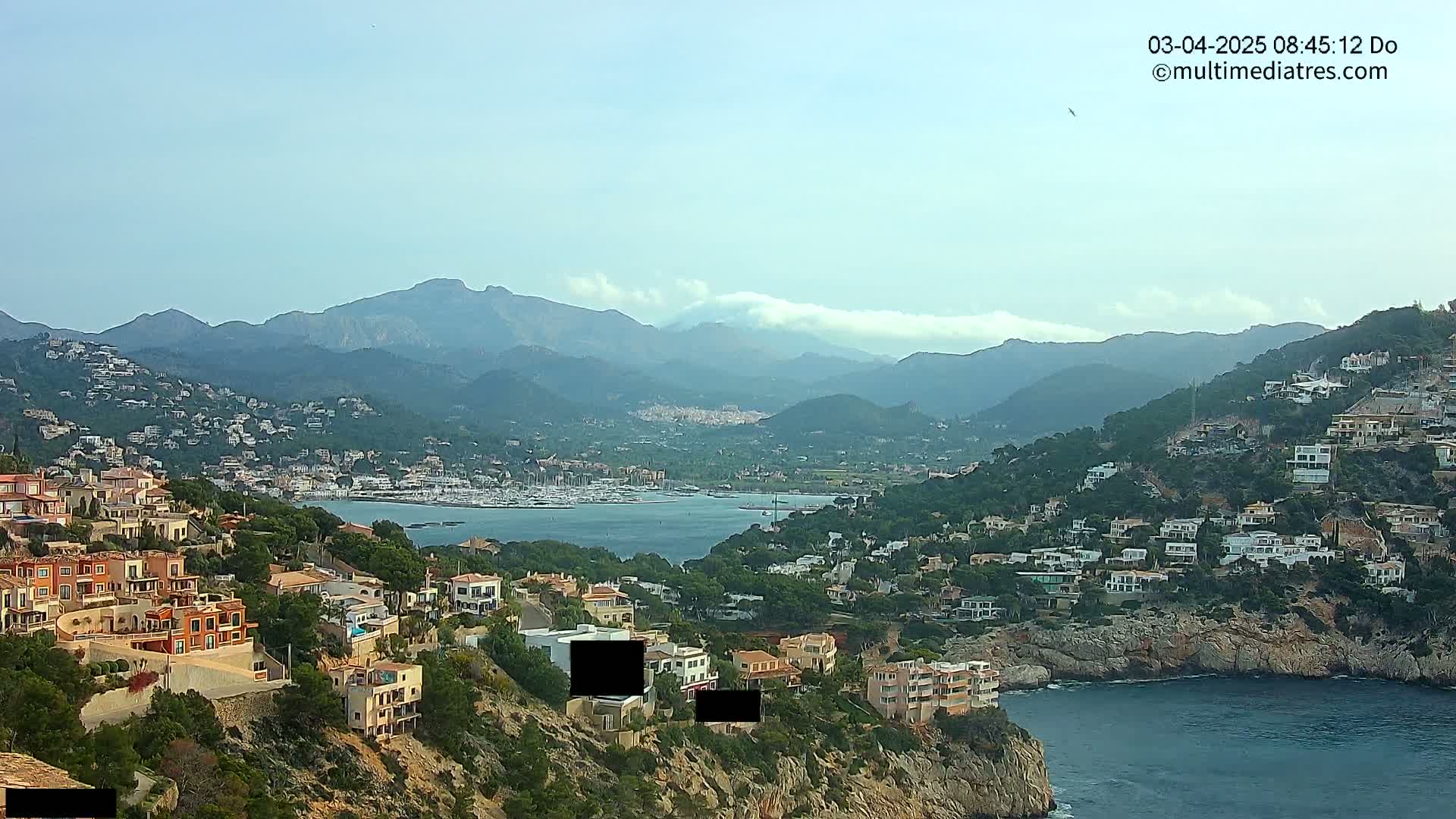 A coastal town nestled in a mountainous region on a partly cloudy day, with a calm bay filled with boats in the background.
