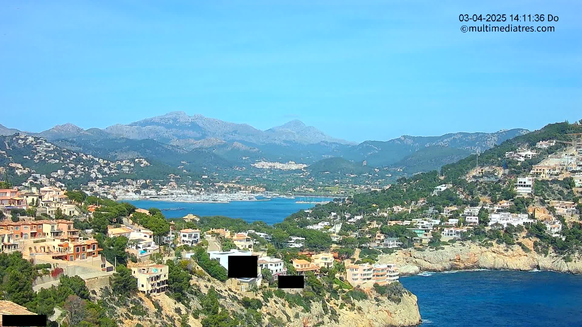 A coastal town nestled in a hillside overlooking a calm bay, with mountains in the background under a clear blue sky.