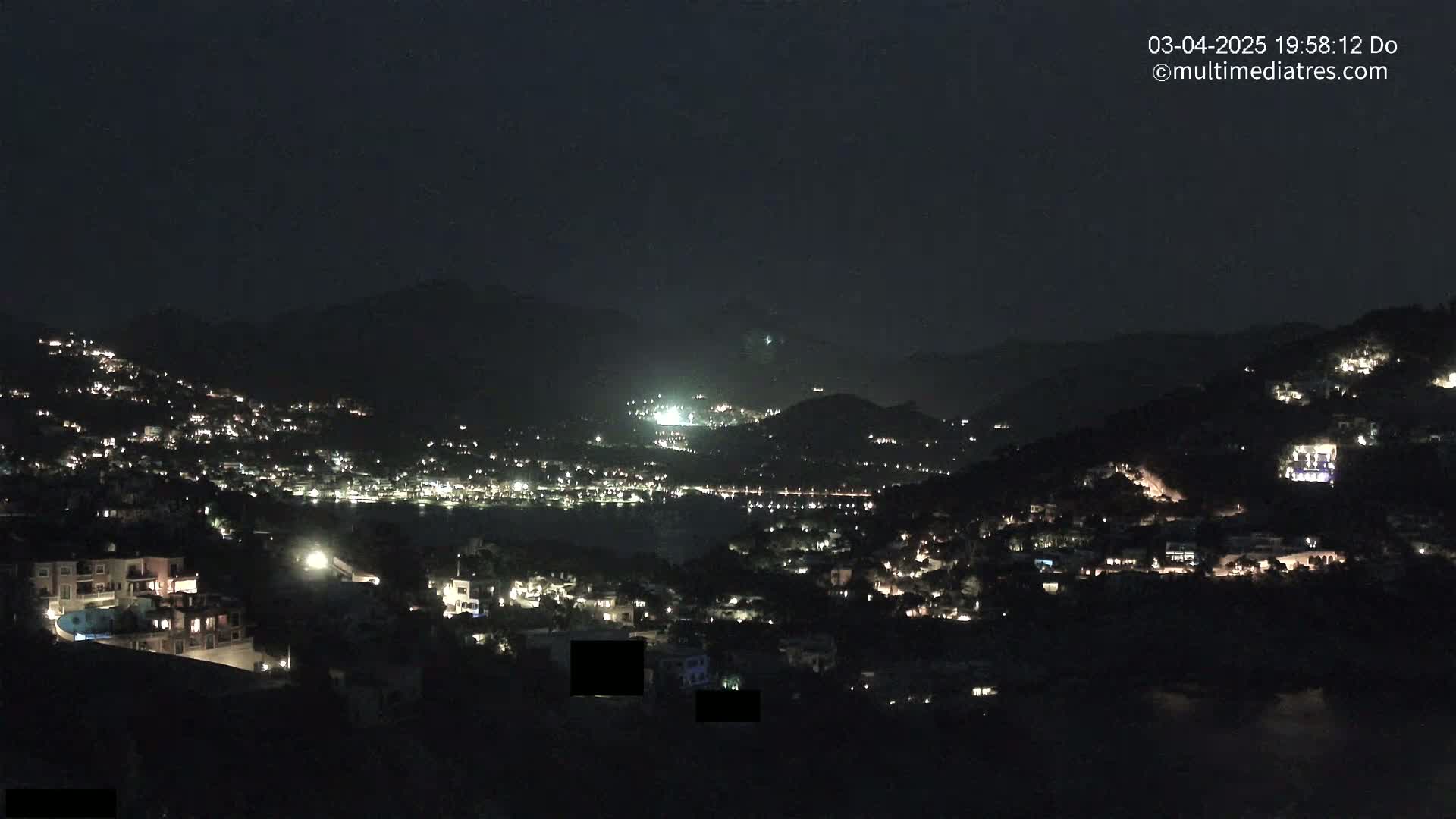 A nighttime view of a hillside town illuminated by scattered lights, with darker hills and a body of water visible in the distance under a clear, dark sky.