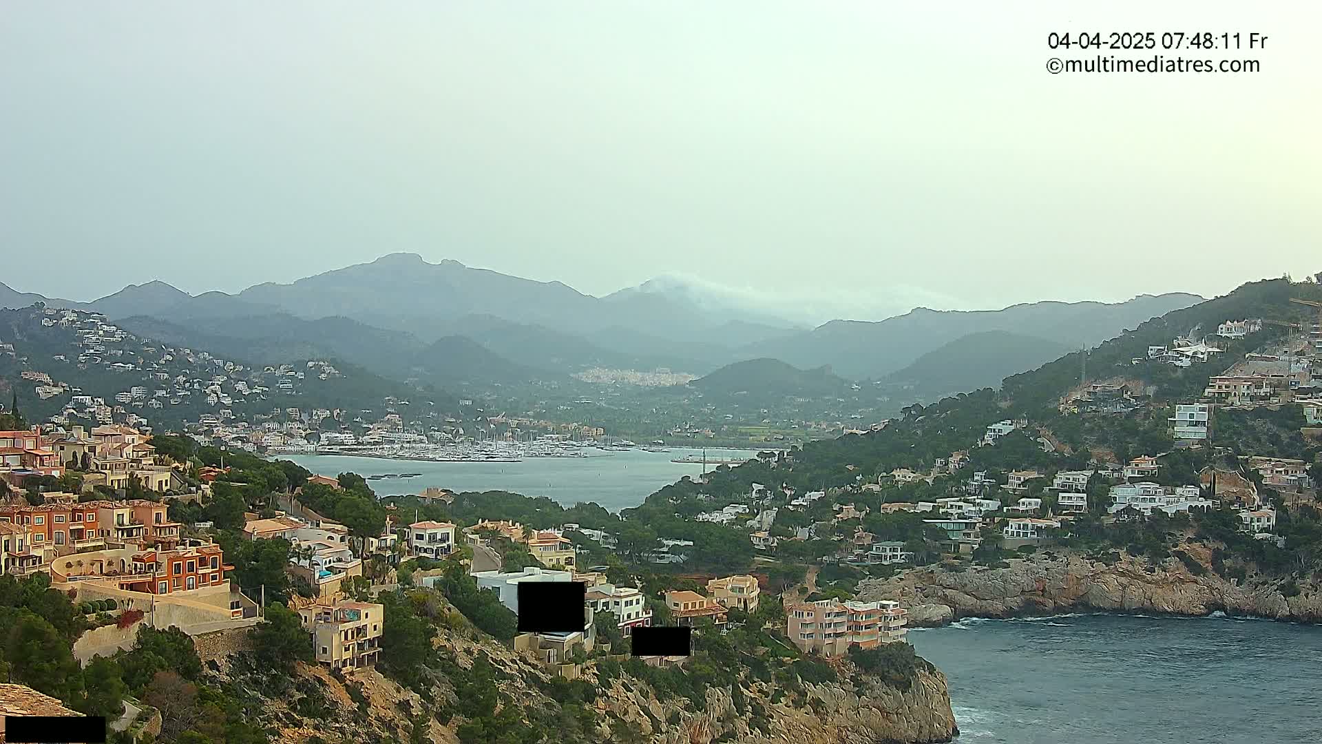 A hazy coastal scene shows a hillside town overlooking a calm bay with many boats, all nestled against a backdrop of mountains.