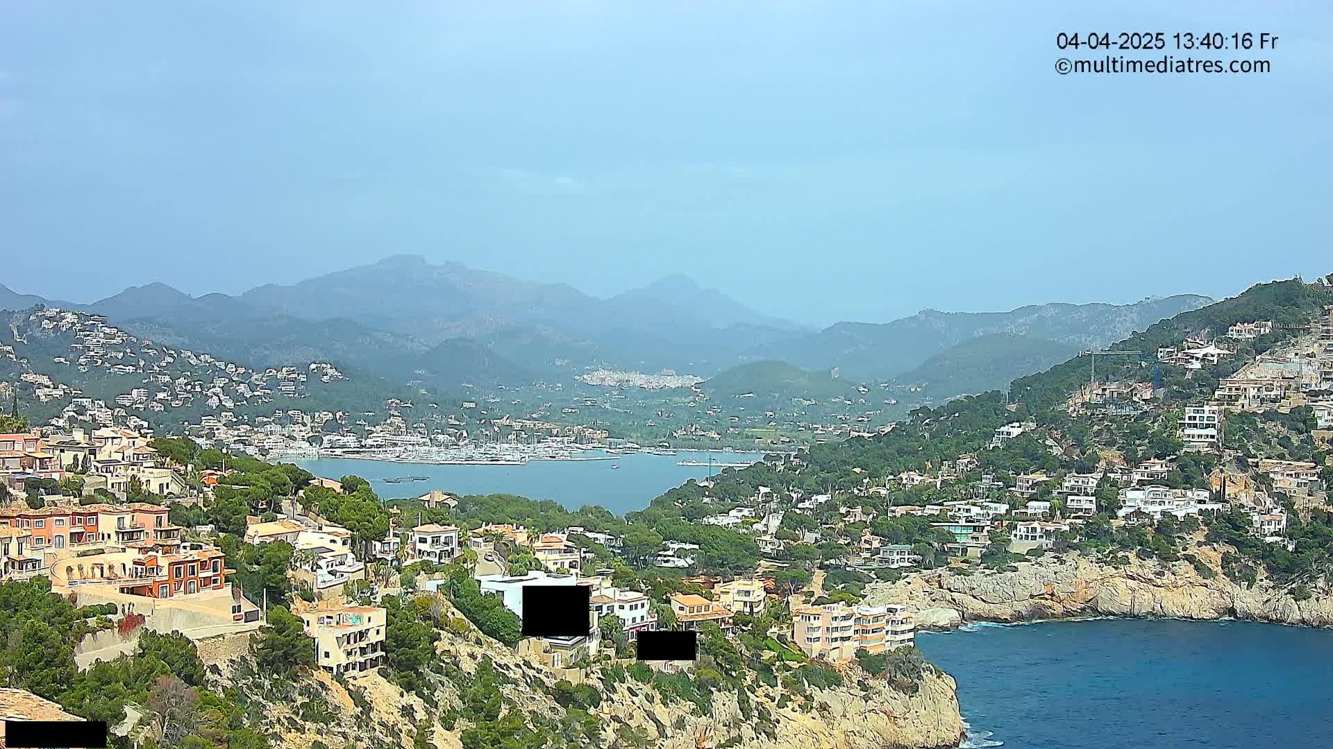 A coastal town nestled on a hillside overlooking a calm bay, with mountains in the background under a clear, hazy sky.
