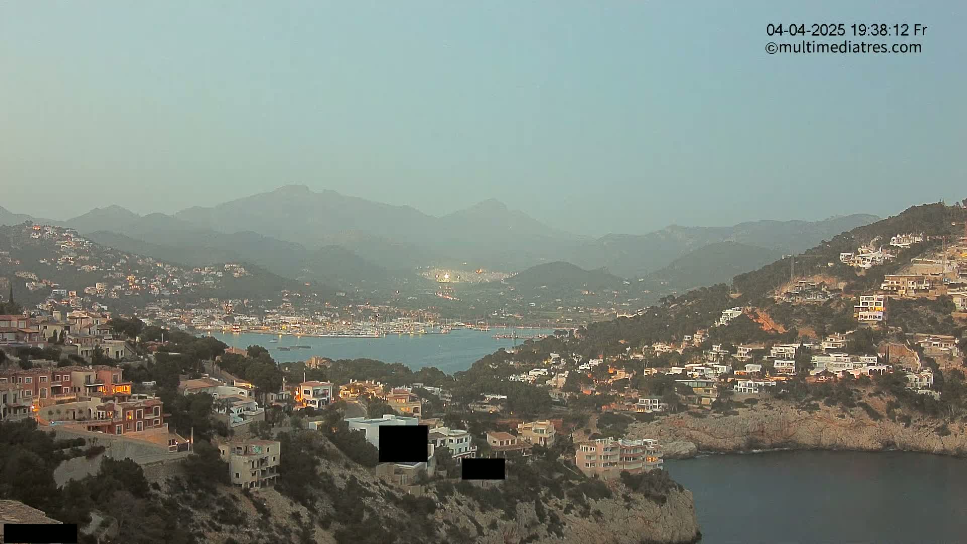 A coastal town nestled on a hillside at dusk, overlooking a calm bay and distant hazy mountains.